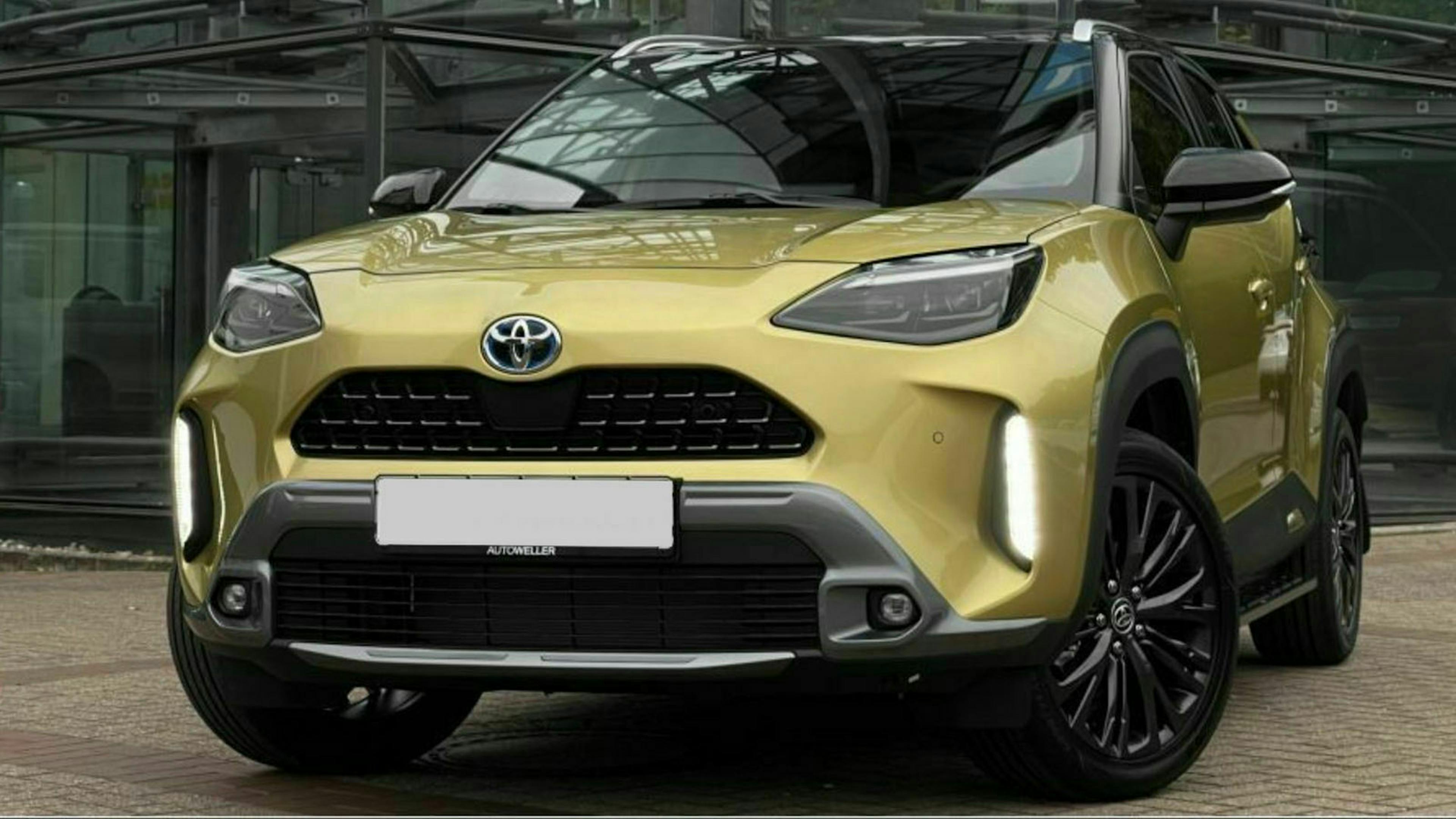 Ein goldener Toyota Yaris Cross steht auf einem Parkplatz vor einem Gebäude mit Fensterfront