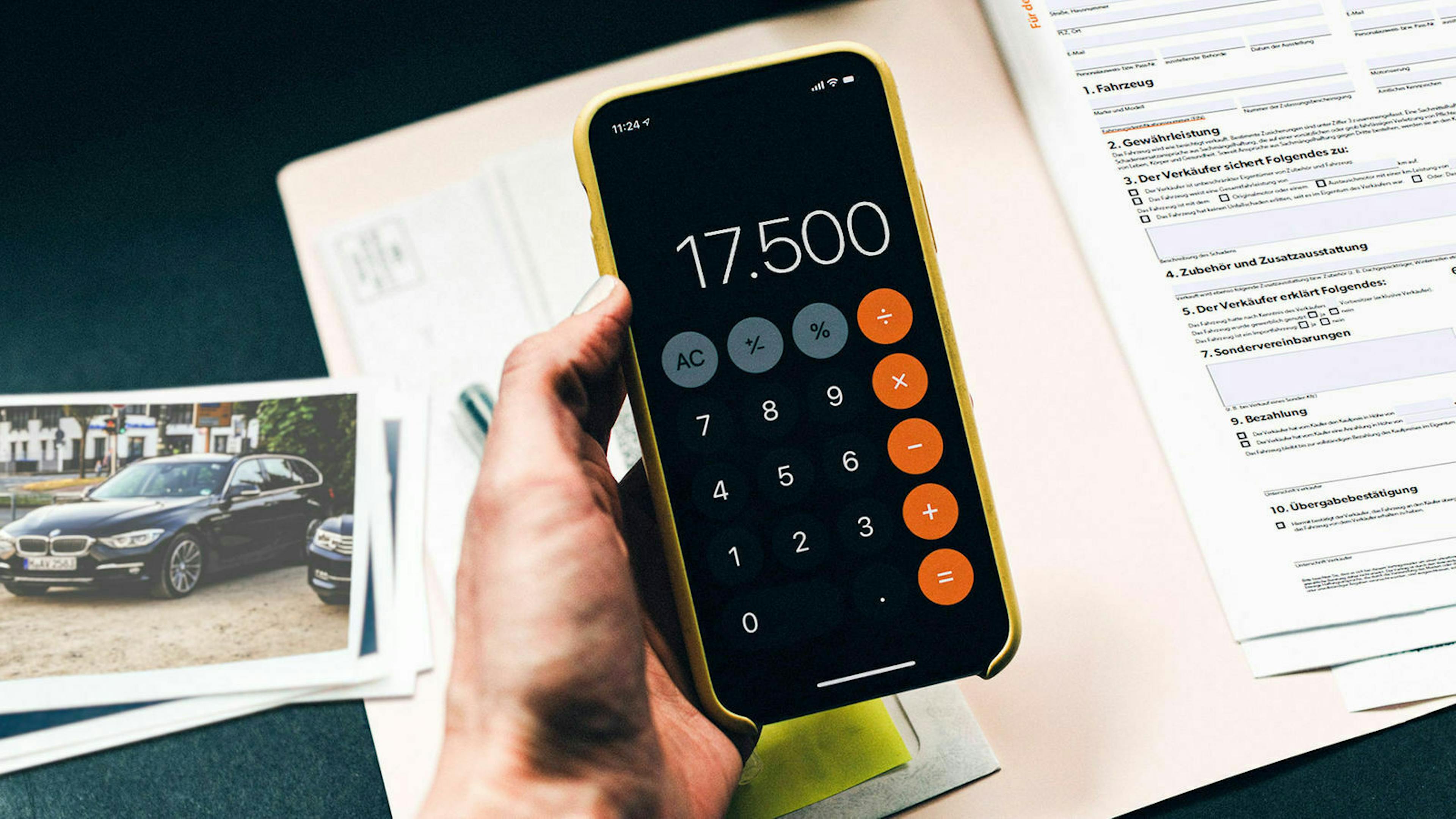 Jemand hält ein Smartphone mit geöffneter Taschenrechner-App in der Hand. Links davon liegt ein Foto von einem Auto.