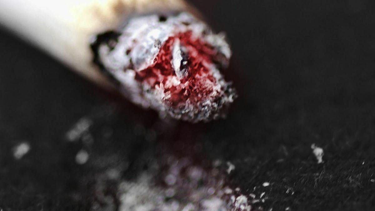 Eine glühende Zigarette liegt auf einem Autoteppich, auf dem sich bereits ein Brandloch bildet.
