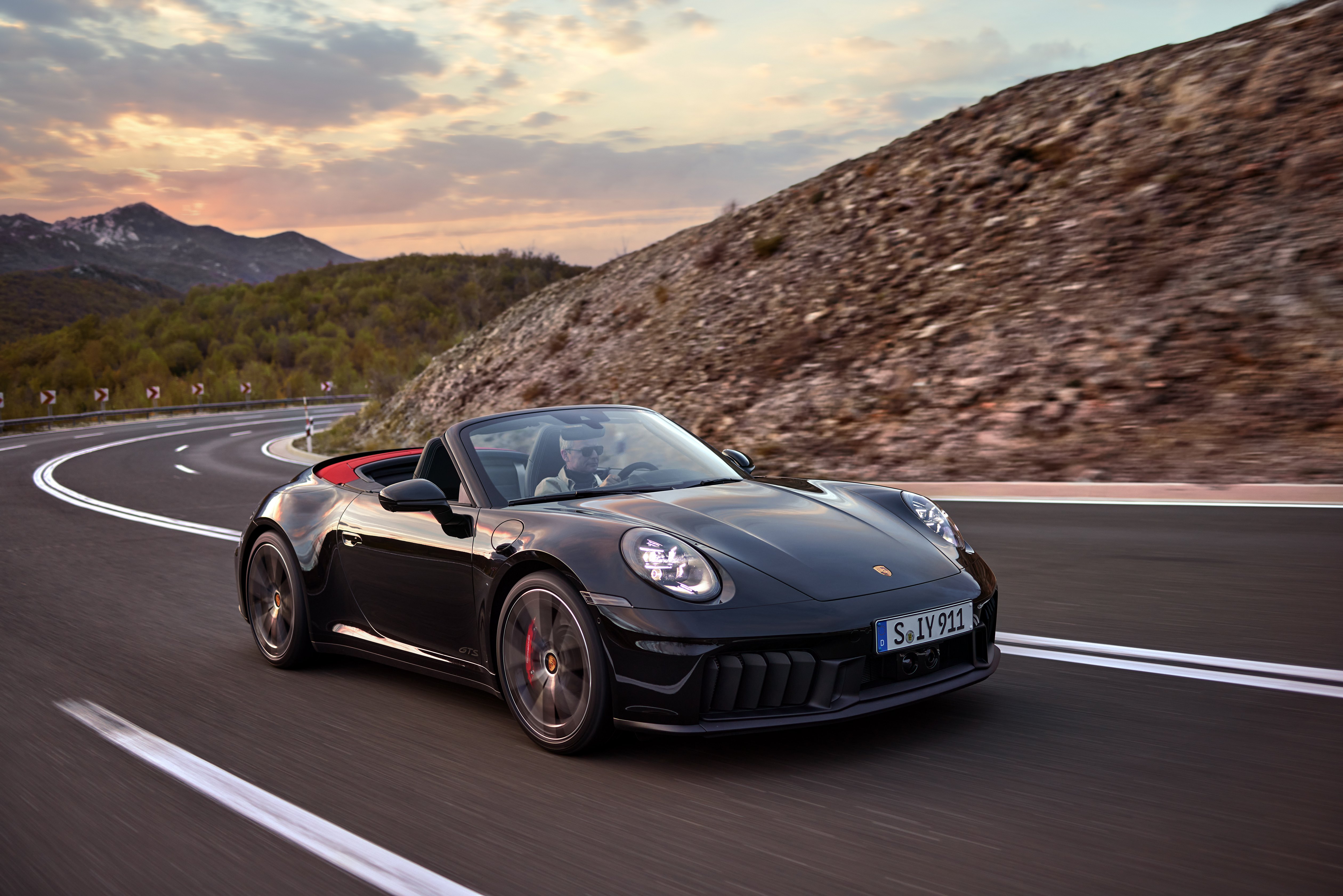Ein schwarzes Porsche 911 Cabriolet fährt im Sonnenuntergang auf einer Landstraße.