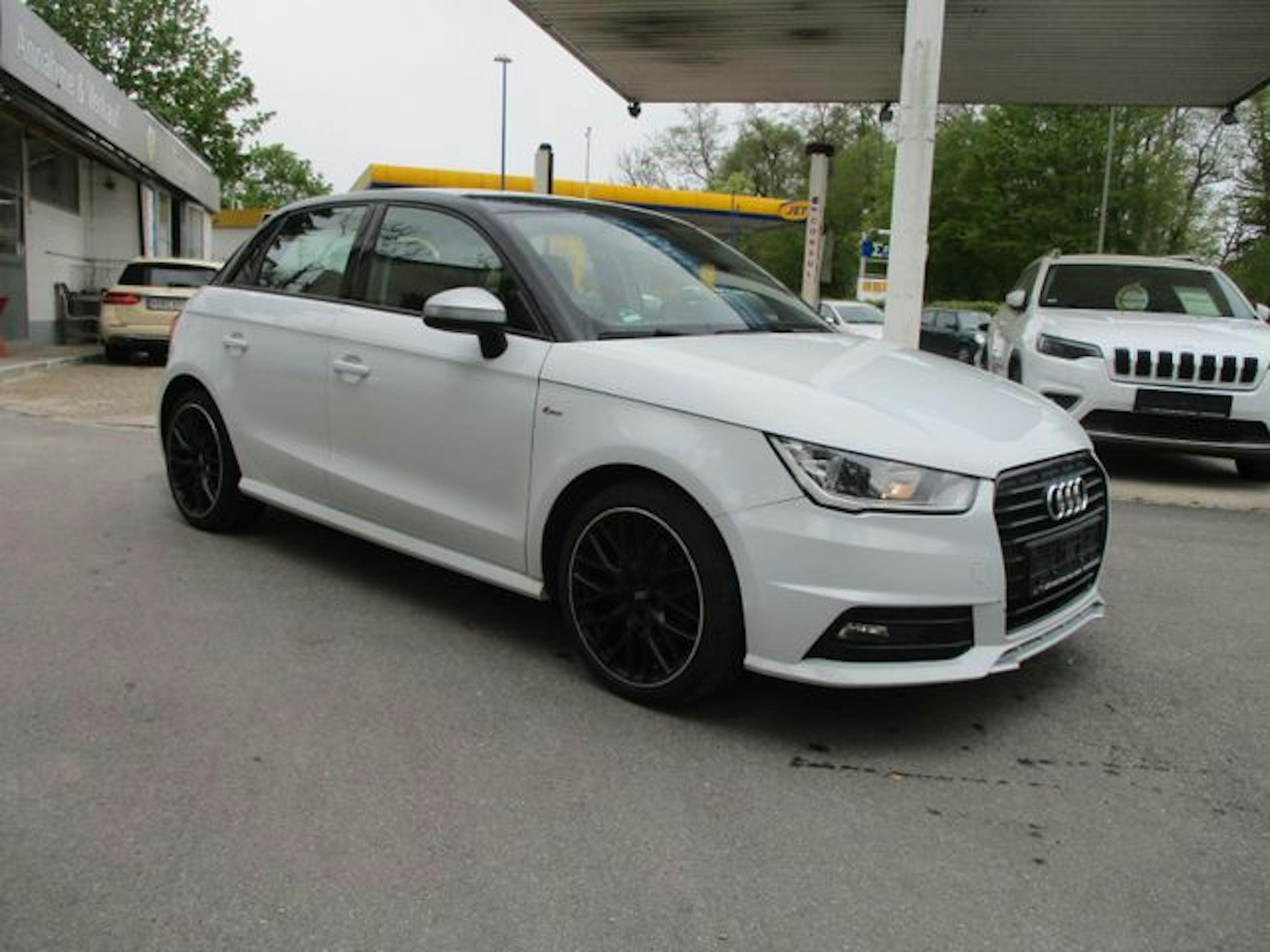 Weißer Audi A1 mit schwarzen Felgen auf einem Parkplatz, umgeben von anderen Autos und einer Tankstelle im Hintergrund.