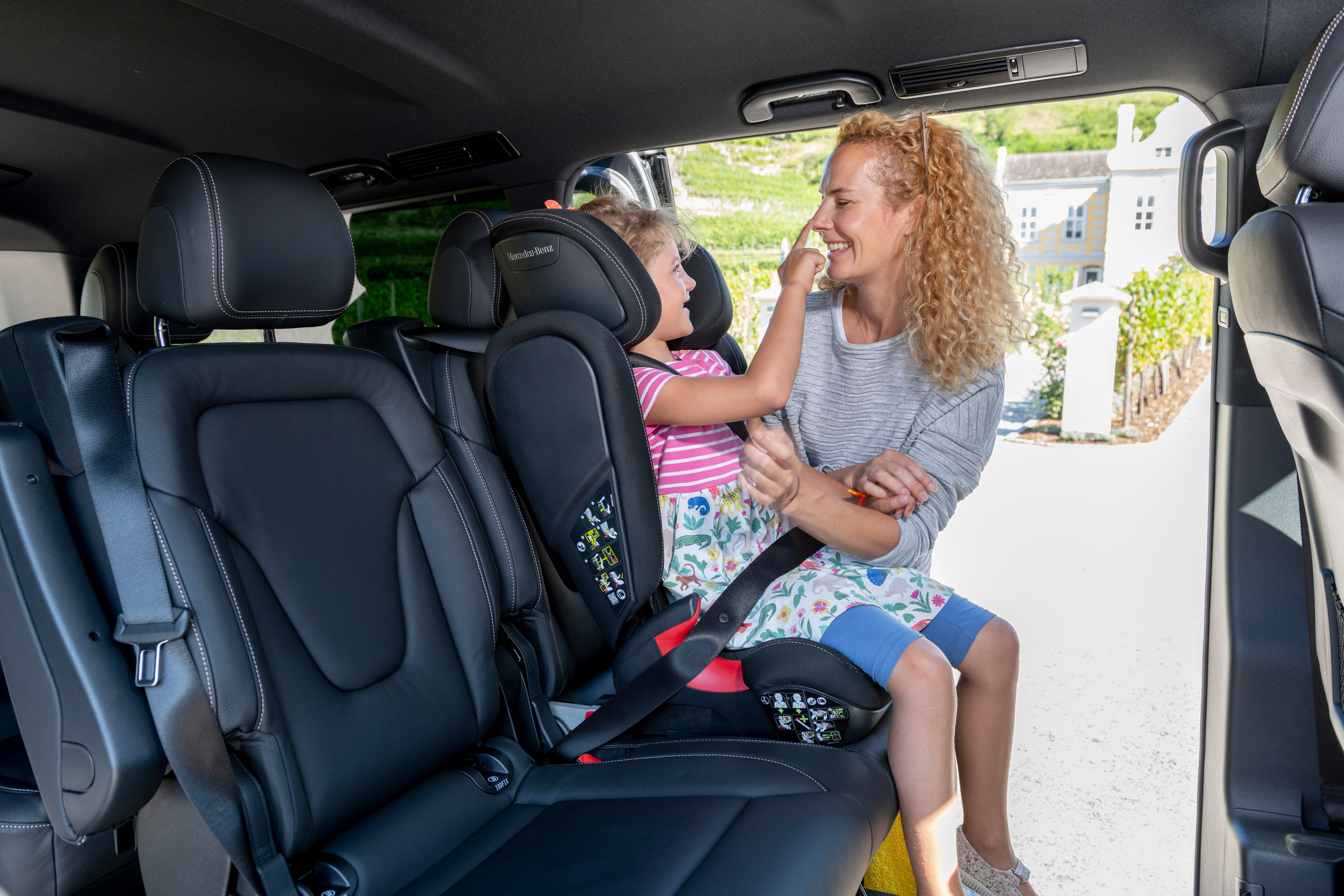 Eine lachende Frau und ein Kind sitzen in einem Auto mit offenen Türen. Das Kind berührt das Gesicht der Frau spielerisch.