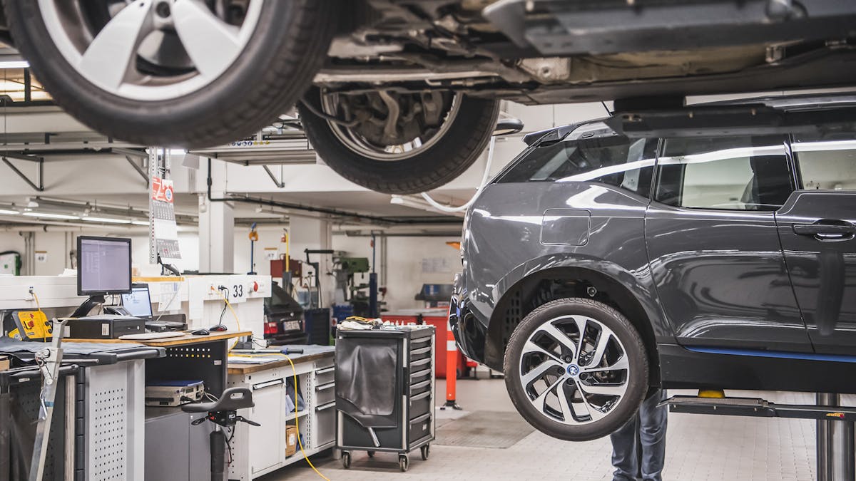 In einer Werkstatt stehen zwei Autos auf einer Hebebühne. Im Hintergrund liegen Werkzeuge auf einer Werkbank.