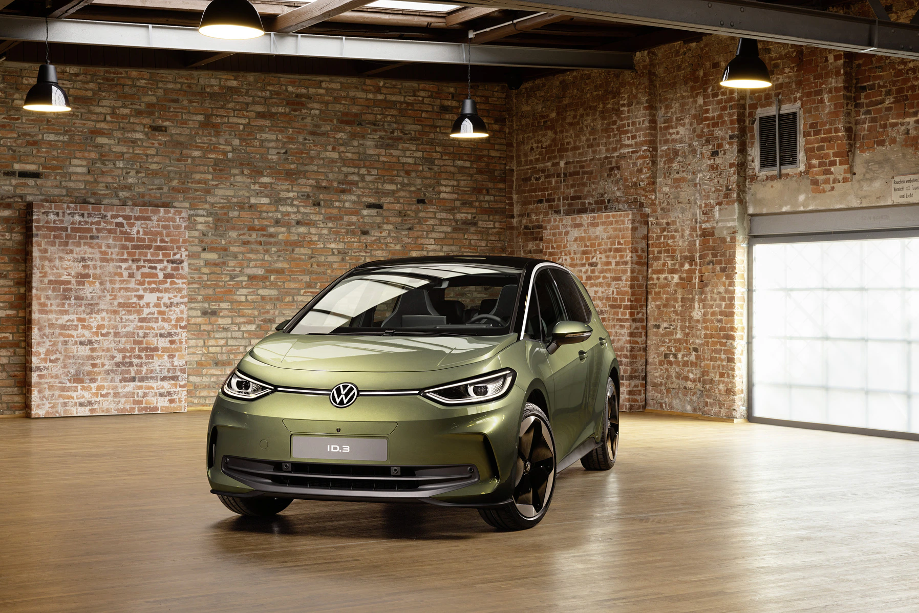 Ein olivgrüner VW ID.3 steht in einem Loft mit Backsteinwänden und Holzboden. Industrielle Hängelampen beleuchten den modernen Elektroauto.