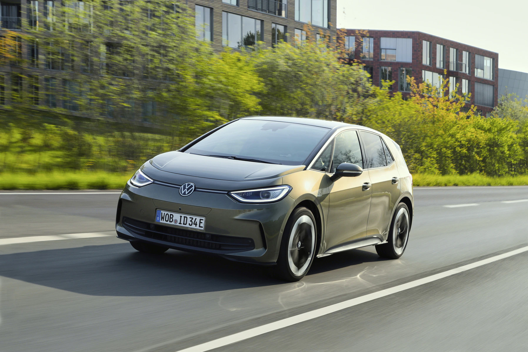 Ein grauer VW ID.3 fährt auf einer Landstraße, umgeben von grüner Natur und modernen Gebäuden im Hintergrund.