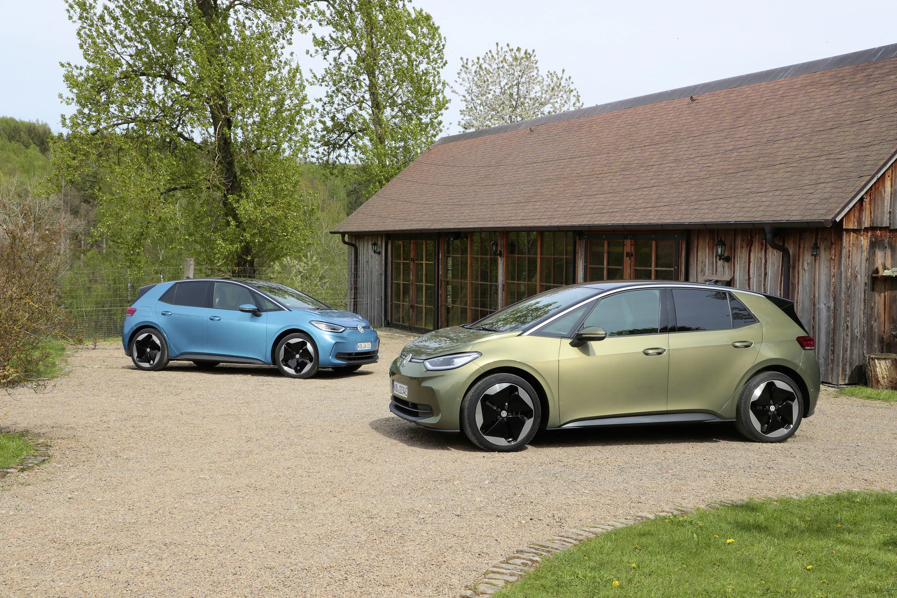 Zwei VW ID.3 Elektroautos in Blau und Grün stehen auf einem Kiesweg vor einer Holzhütte mit Bäumen im Hintergrund.