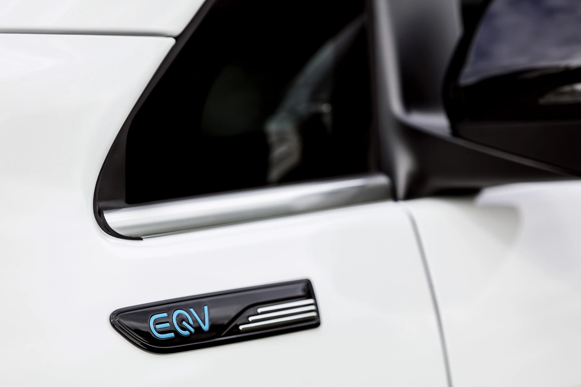 Nahaufnahme eines weißen Mercedes-Benz EQV mit Fokus auf das blaue "EQV"-Emblem an der Seite nahe dem Seitenspiegel.