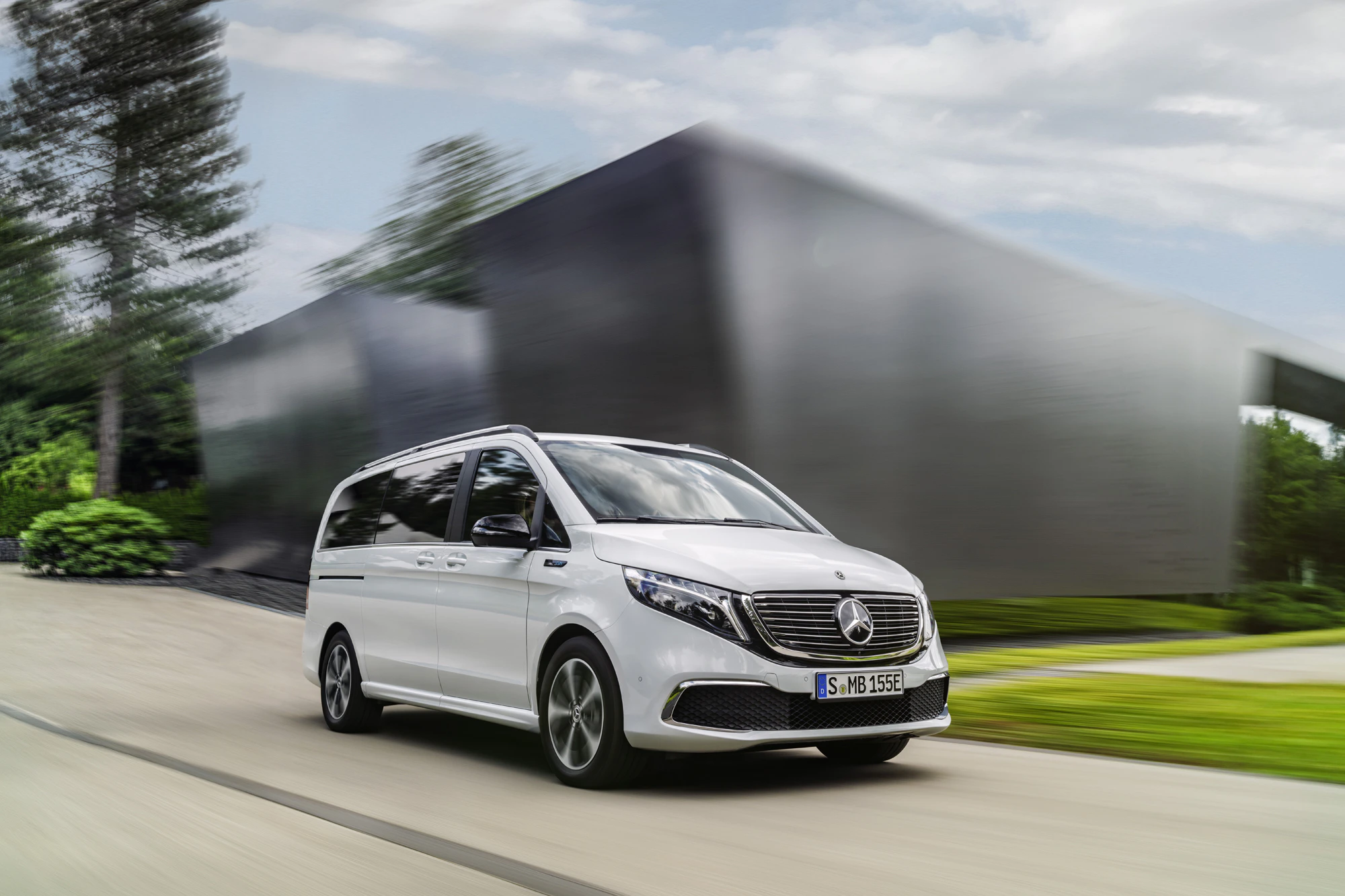Ein weißer Mercedes-Benz EQV fährt dynamisch auf einer Straße, mit moderner Architektur und grüner Landschaft im Hintergrund.