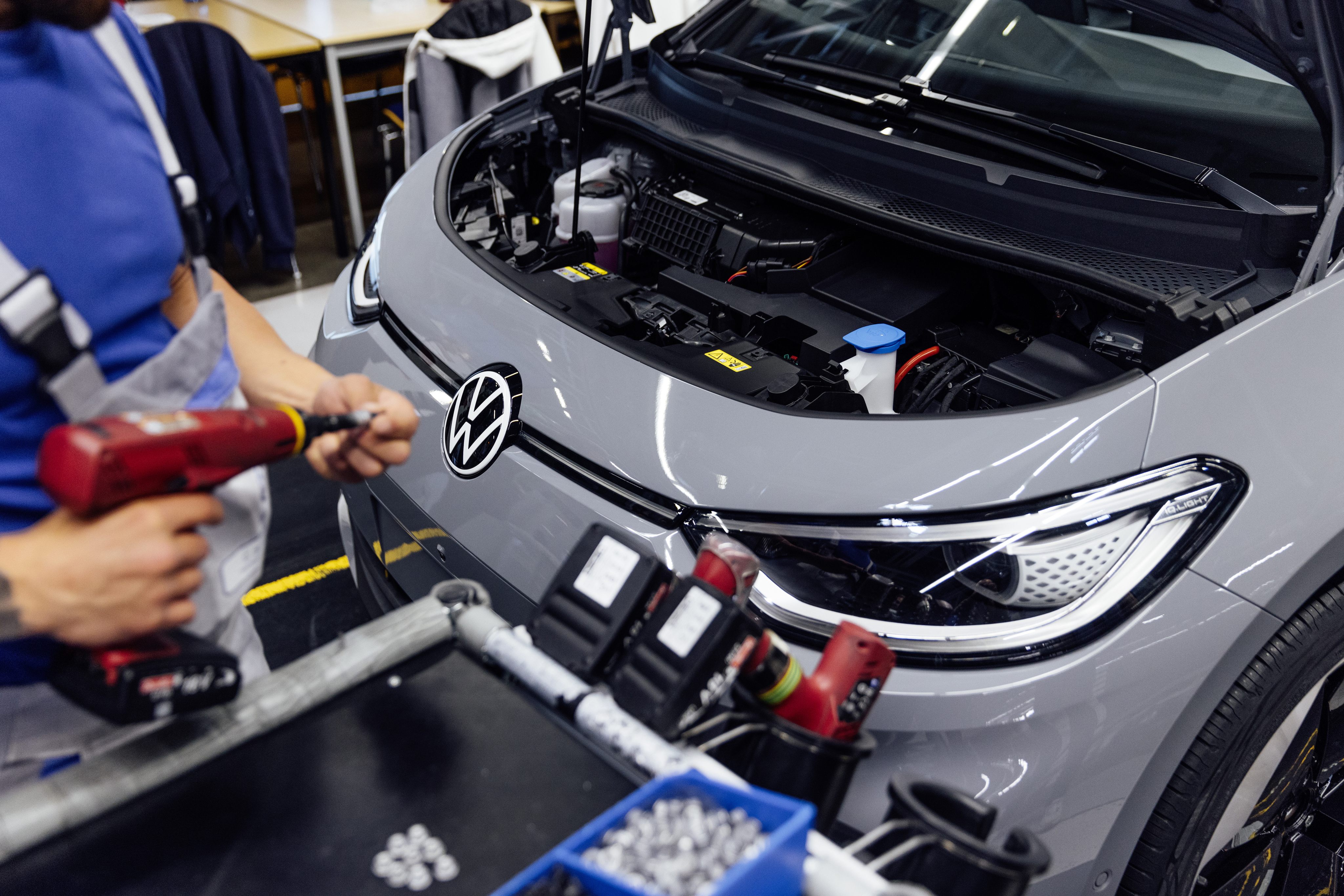Arbeiter montiert ein graues VW-Elektroauto in einer Fabrik, Motorhaube offen, Werkzeuge und Bauteile auf einem Tisch im Vordergrund.