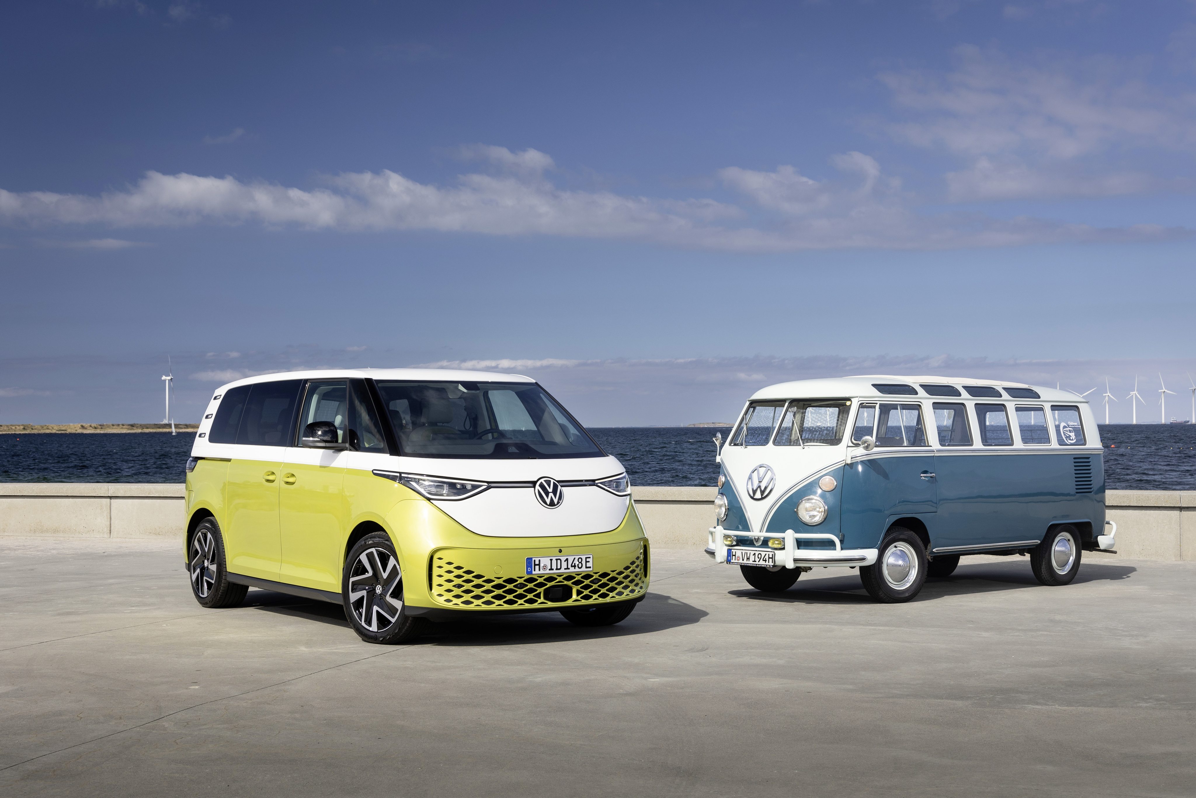 A yellow and white VW ID.Buzz and a blue and white VW Bulli are parked in a parking lot by the sea