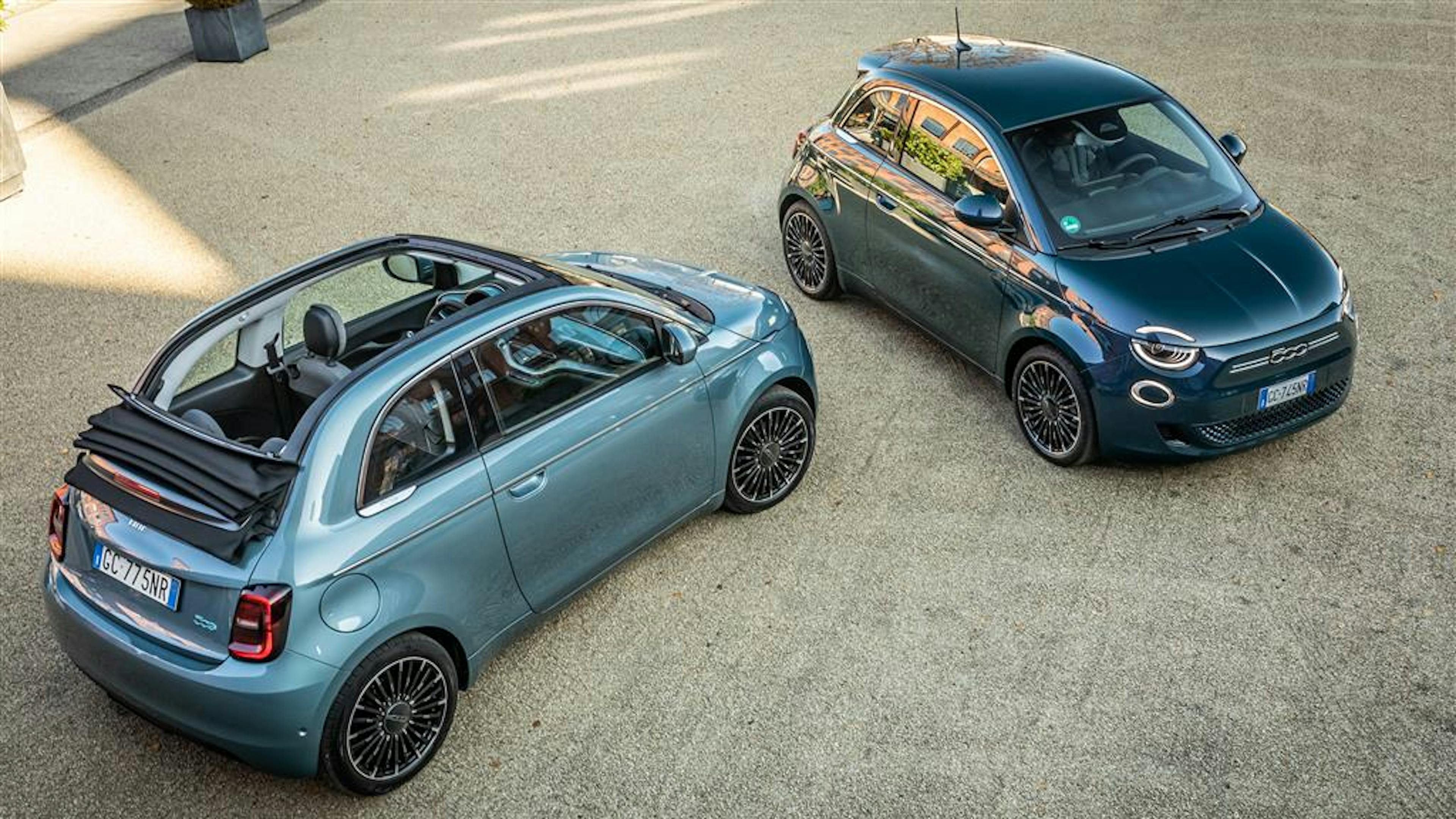 A gray Fiat 500 electric convertible and black Fiat 500 electric are parked on a gravel area.