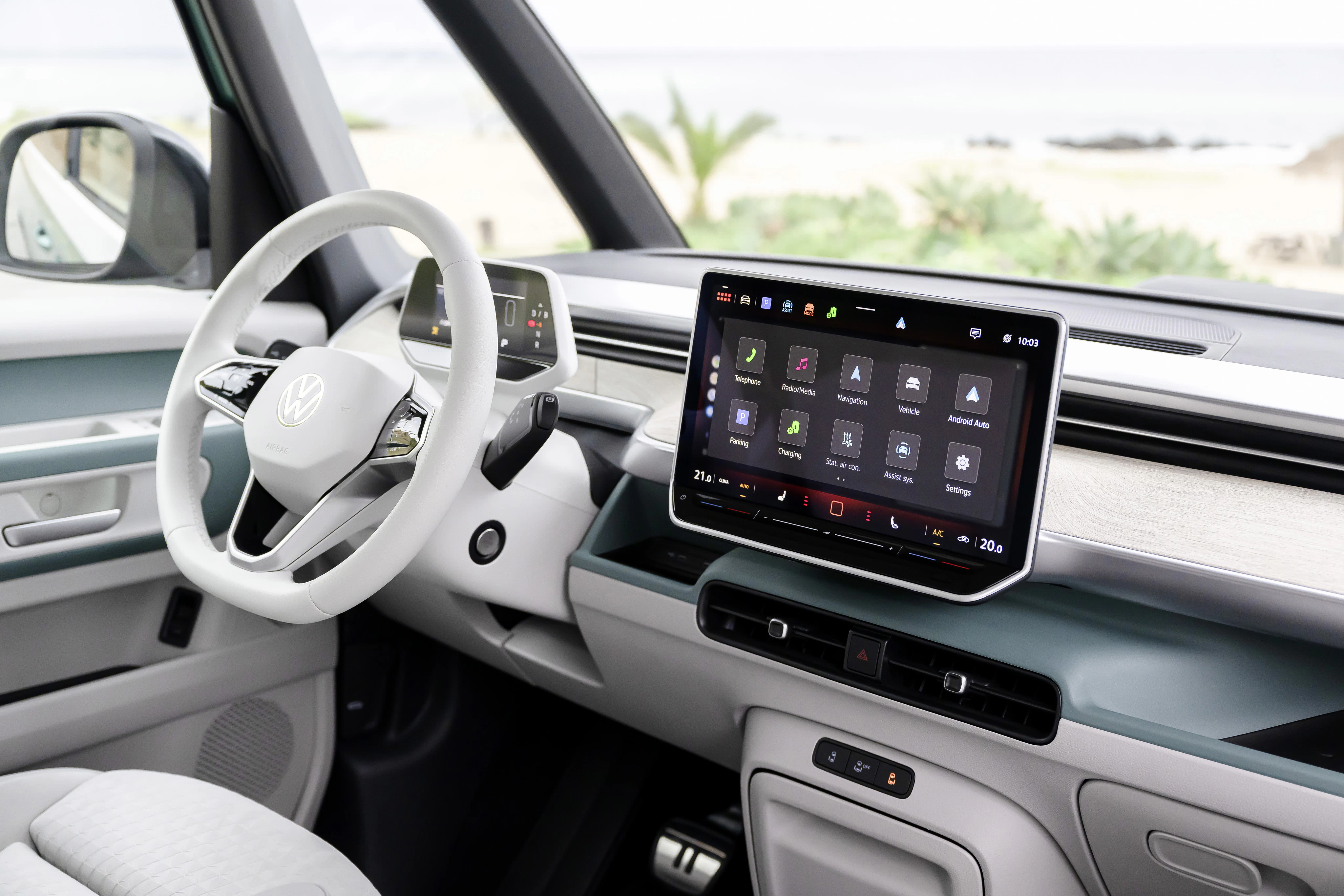 Modernes VW-Auto-Cockpit mit großem Touchscreen, digitalem Display und weißem Lenkrad, mit Blick auf einen Strand durch die Frontscheibe.