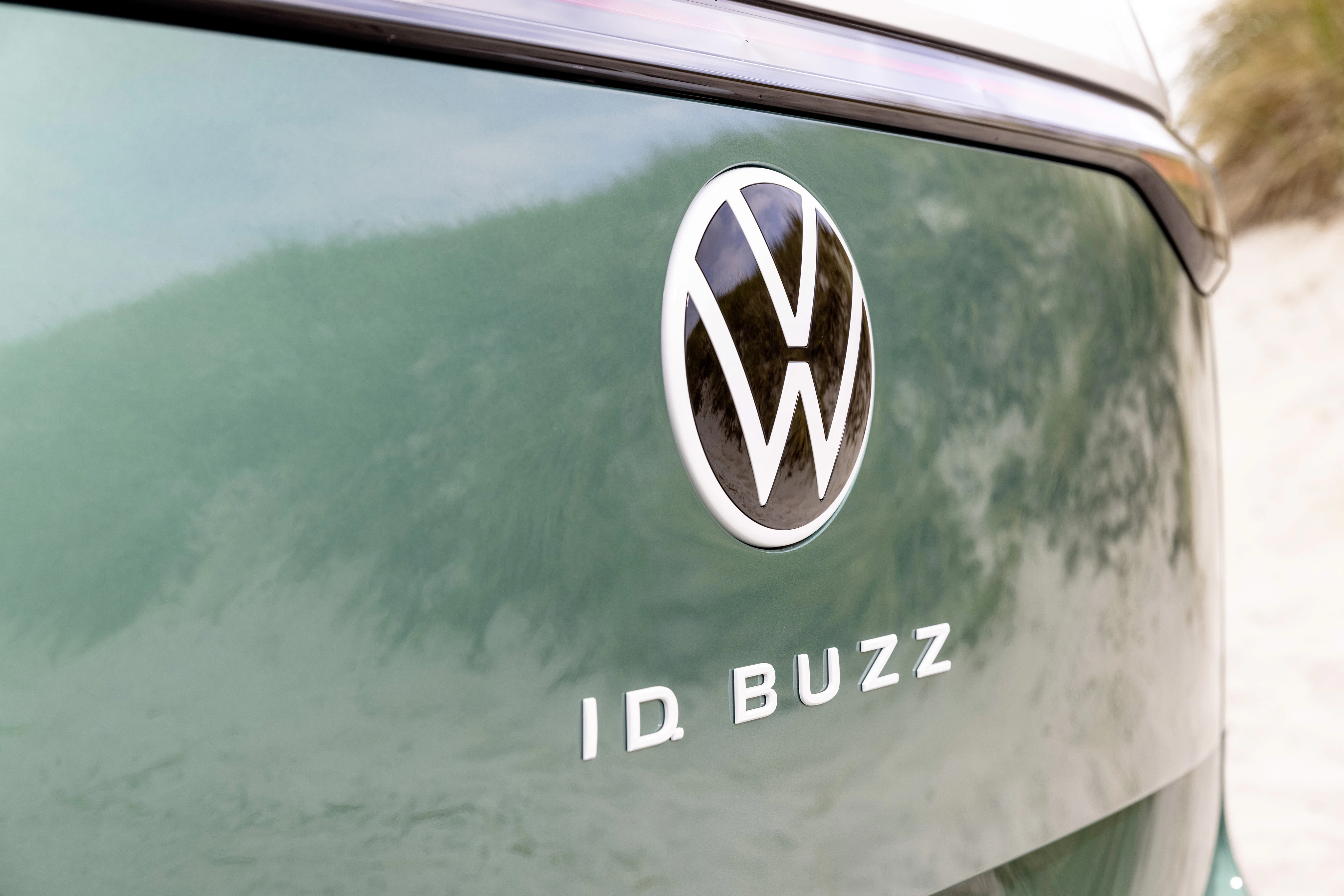 Nahaufnahme des VW-Logos und Schriftzugs „ID. Buzz“ auf der glänzenden Rückseite eines grünen Elektro-Vans vor sandiger Dünenlandschaft.