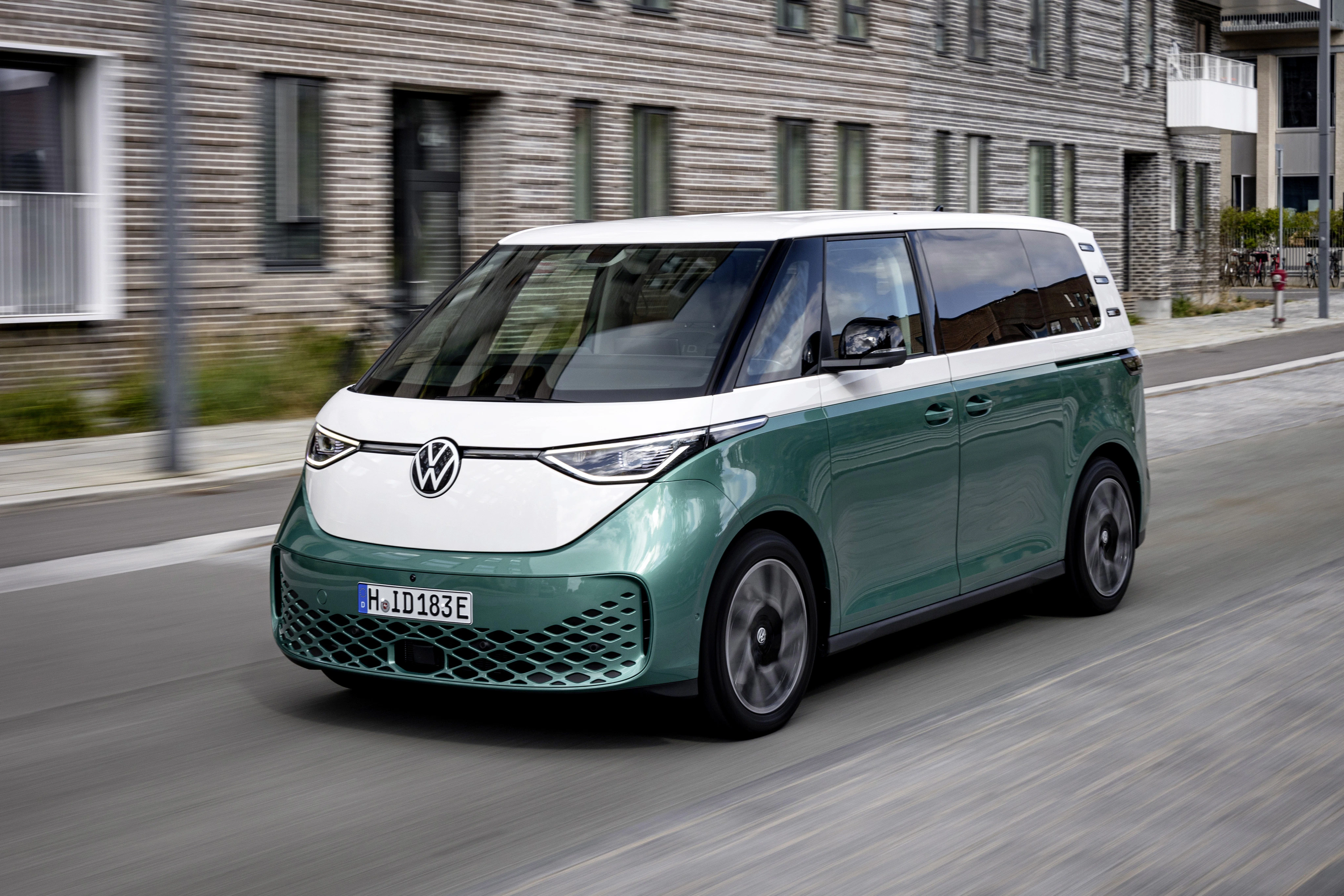 Ein grüner VW ID. Buzz fährt auf einer Straße vor einem modernen Backsteingebäude.