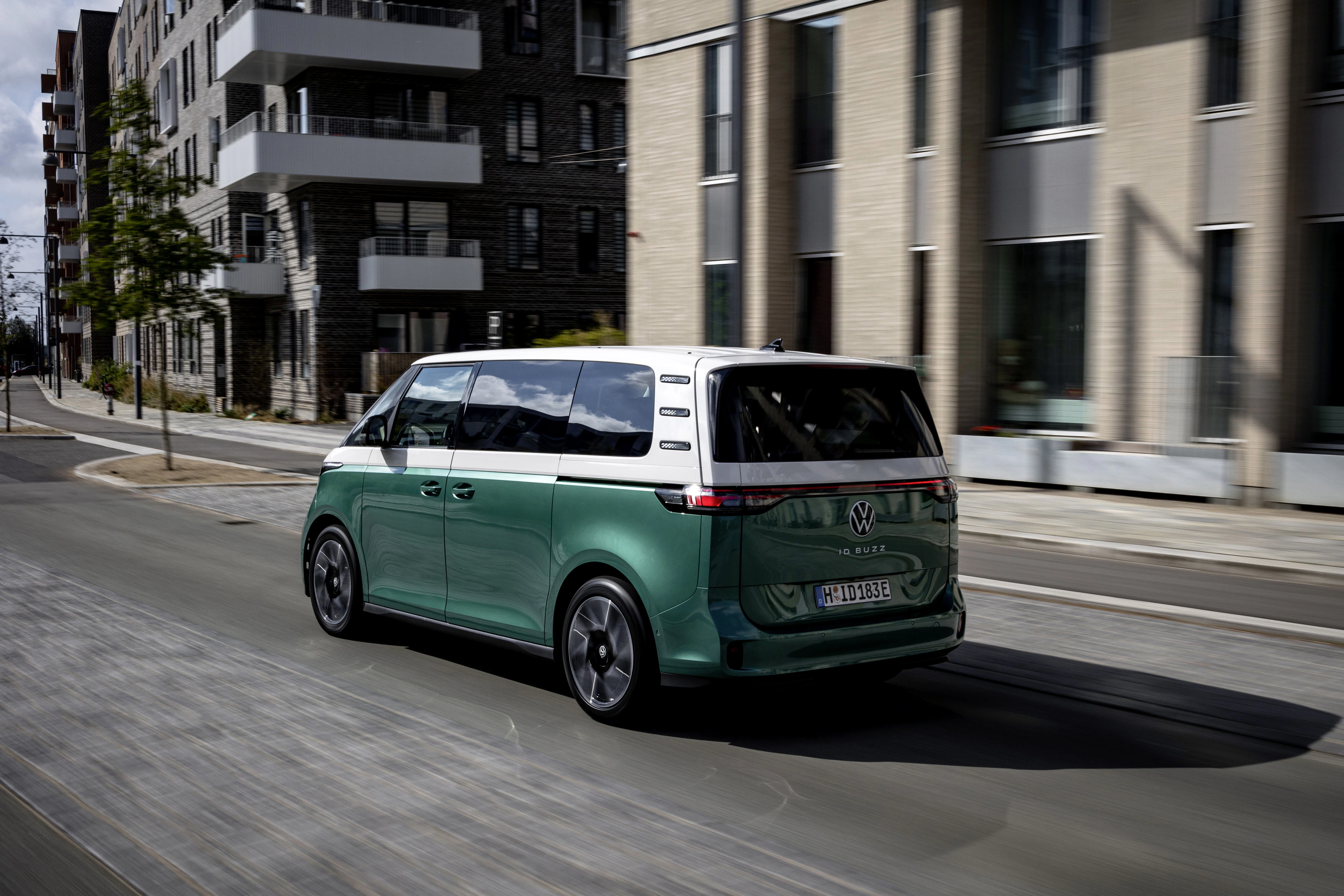 Grüner VW ID. Buzz fährt auf einer städtischen Straße mit modernen Wohngebäuden im Hintergrund. Elektrischer Retro-Van in Bewegung.