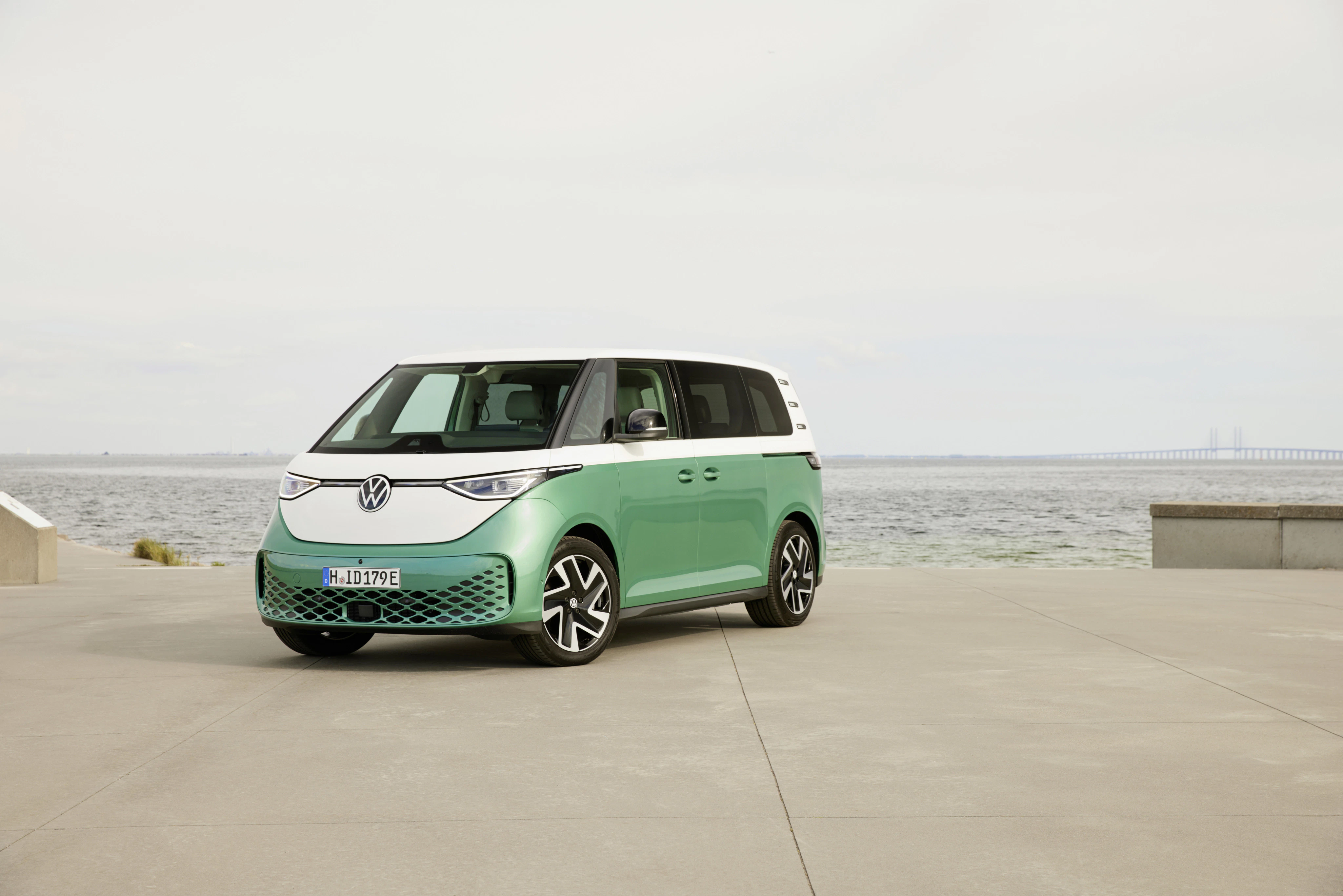 Ein zweifarbiger VW ID. Buzz steht auf einer Betonfläche nahe dem Meer, mit einer Brücke im Hintergrund.