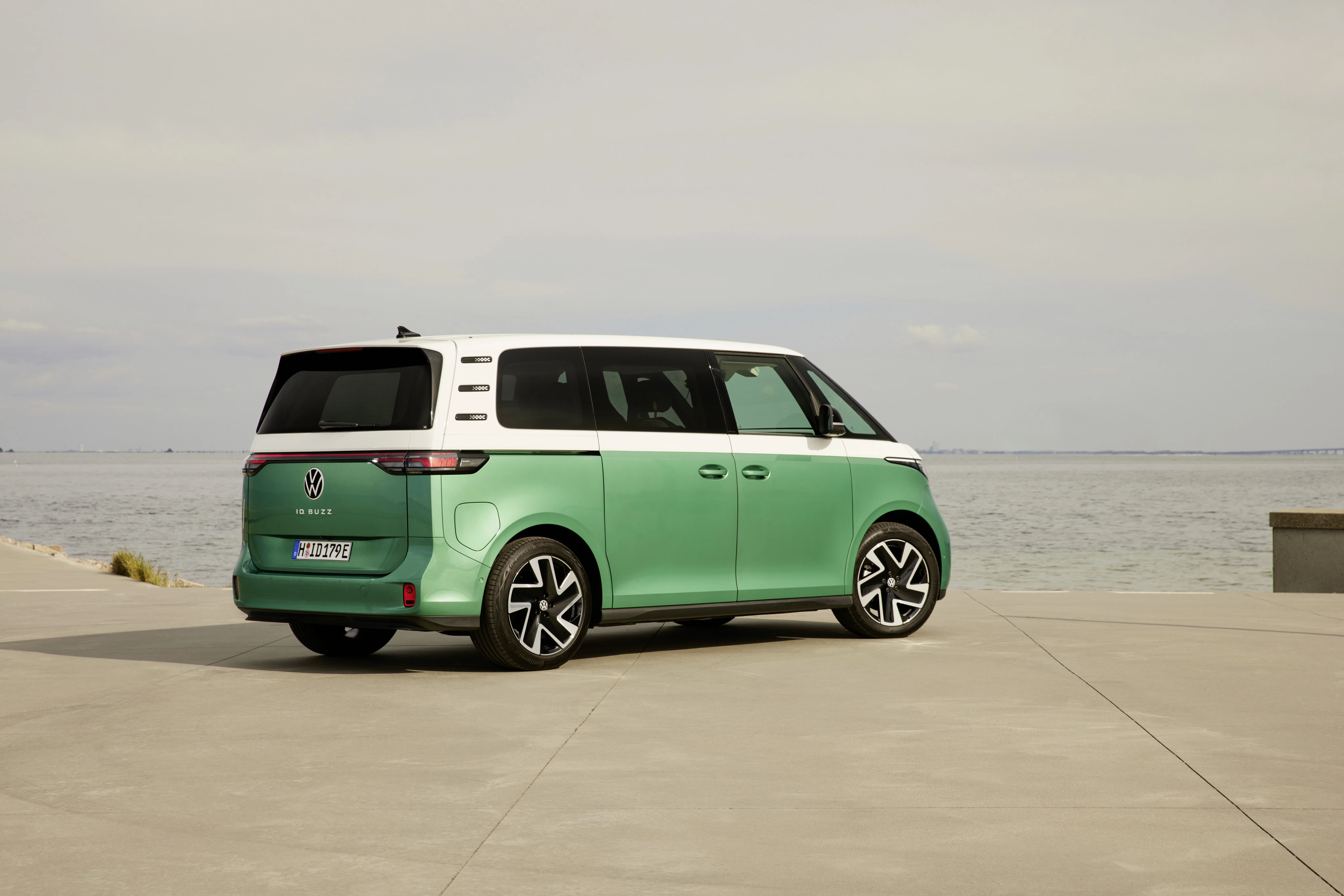Grüner VW ID. Buzz mit weißem Dach steht auf einer Betonfläche am Wasser, Blick aufs Meer unter bewölktem Himmel.