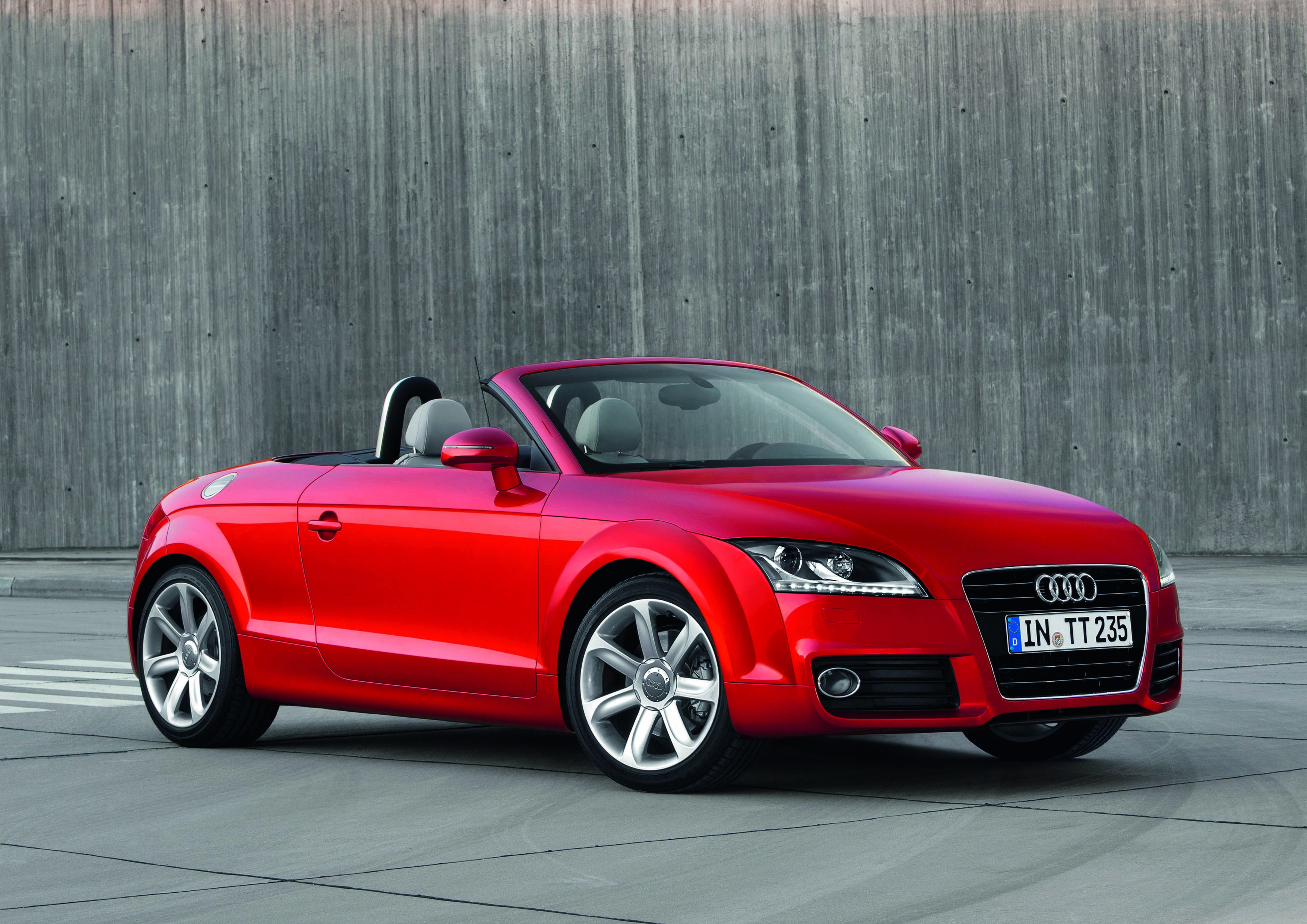 Ein roter Audi TT Roadster mit offenem Verdeck parkt vor einer grauen Holzwand.