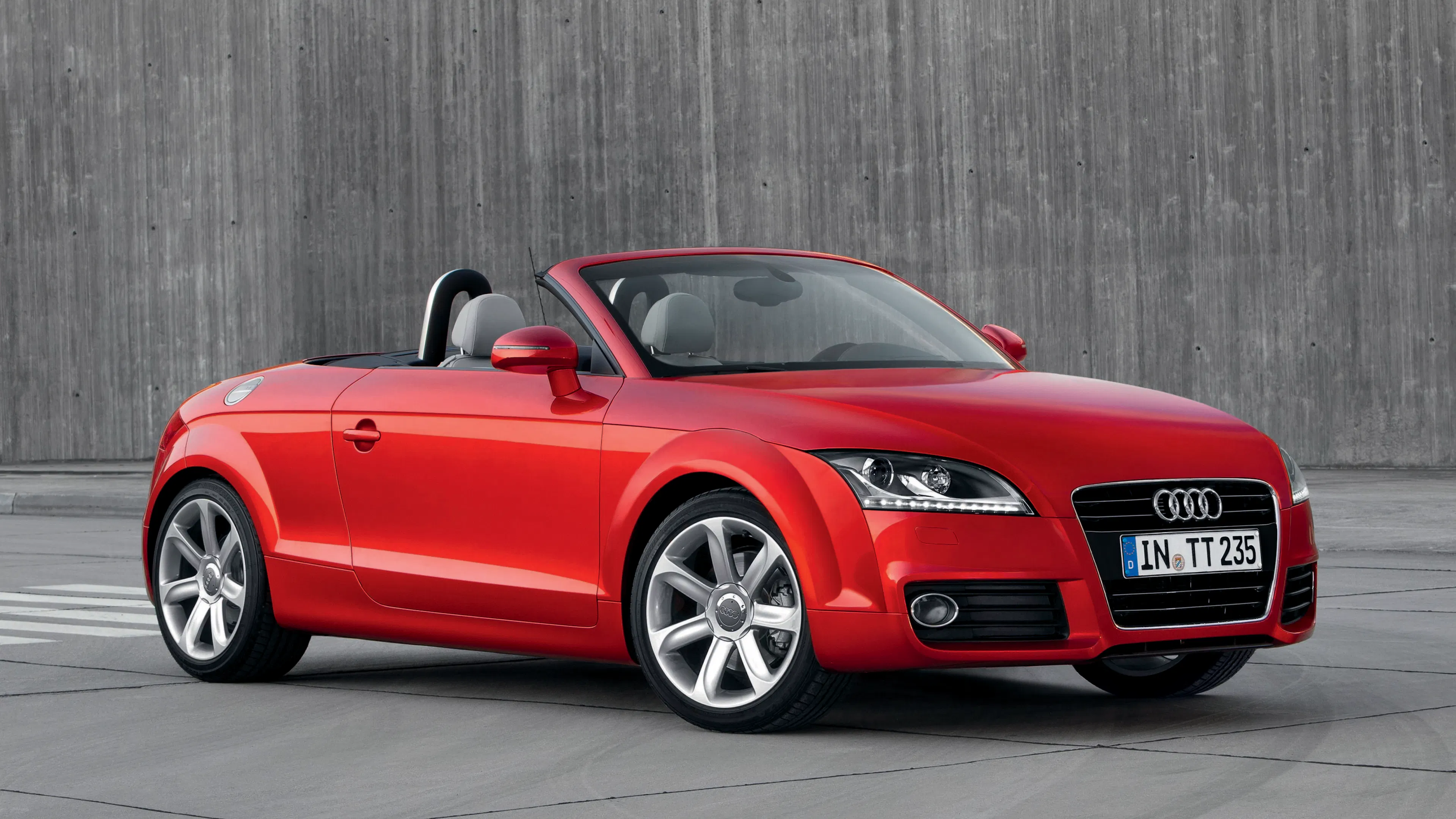 Ein roter Audi TT Roadster mit offenem Verdeck parkt vor einer grauen Holzwand.