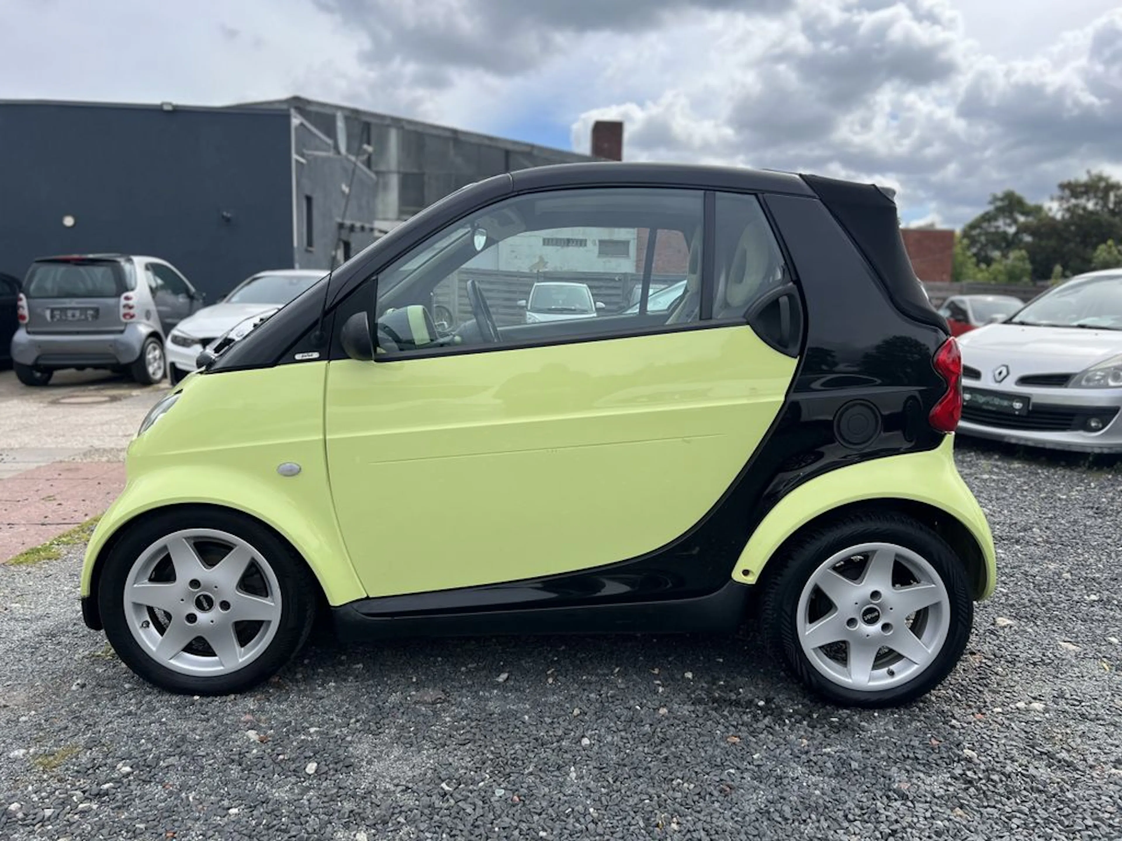Ein gelb-schwarzes Smart Fortwo Cabrio parkt auf einer Schotterfläche.