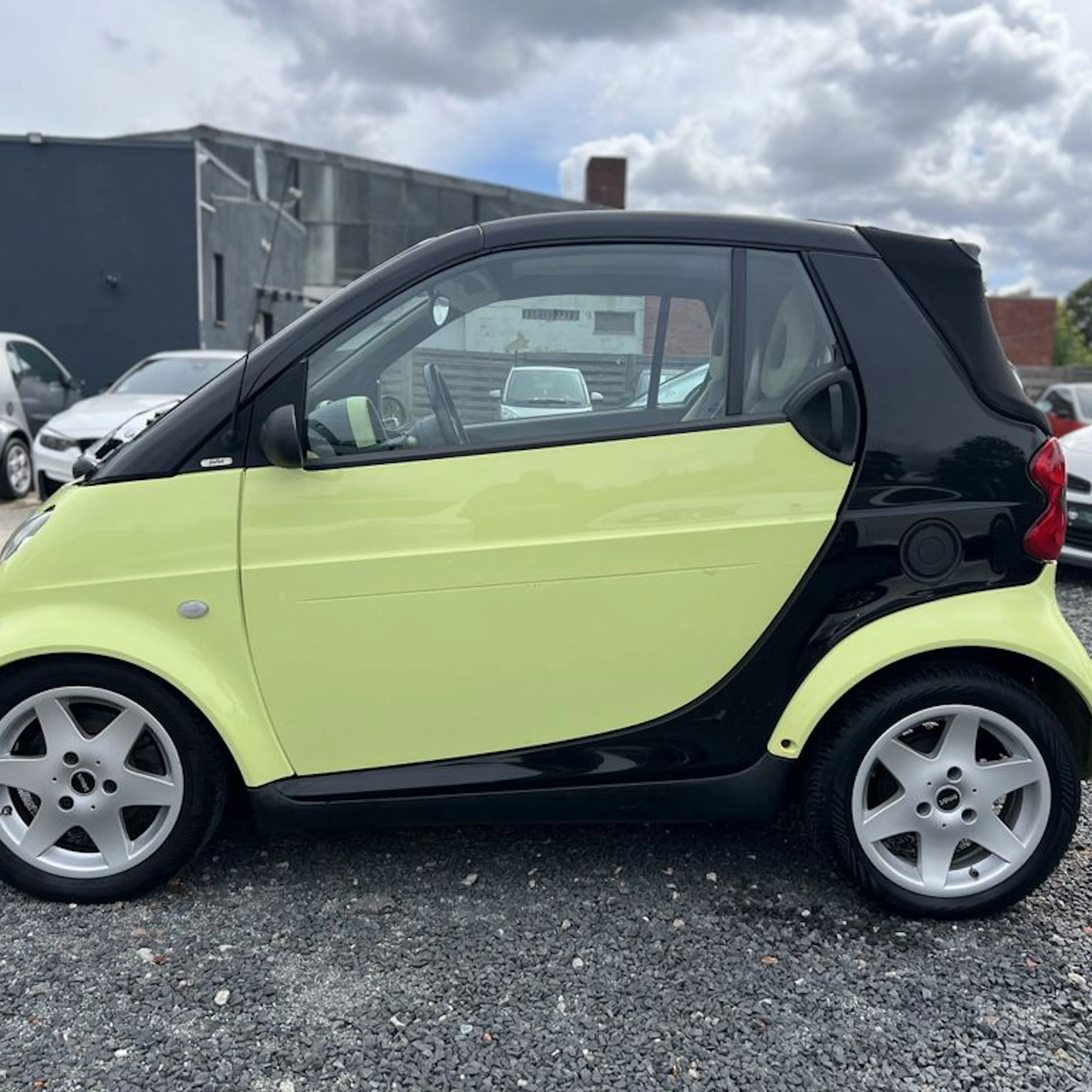Ein gelb-schwarzes Smart Fortwo Cabrio parkt auf einer Schotterfläche.