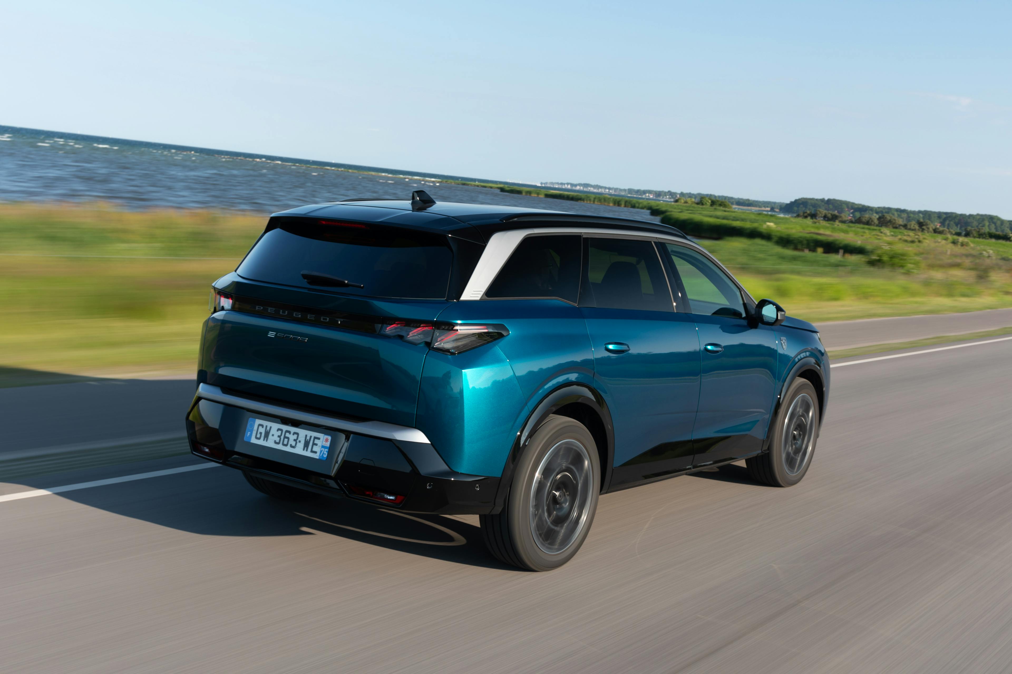 Ein blau-metallic-farbener Peugeot E-5008 fährt auf einer Küstenstraße, im Hintergrund ist das Meer zu sehen.