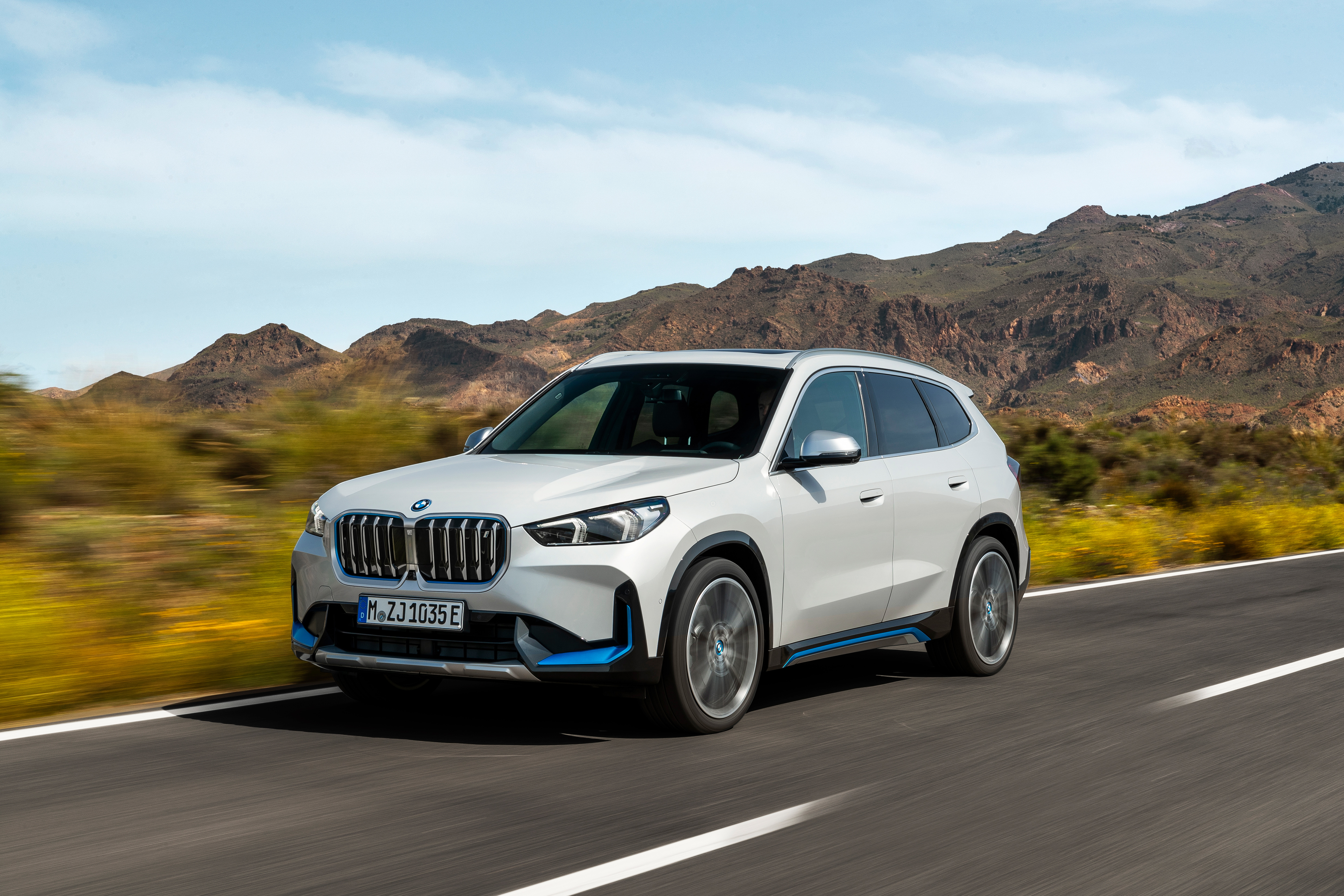 Weißer BMW-SUV fährt auf einer Landstraße durch eine trockene, hügelige Landschaft unter blauem Himmel.