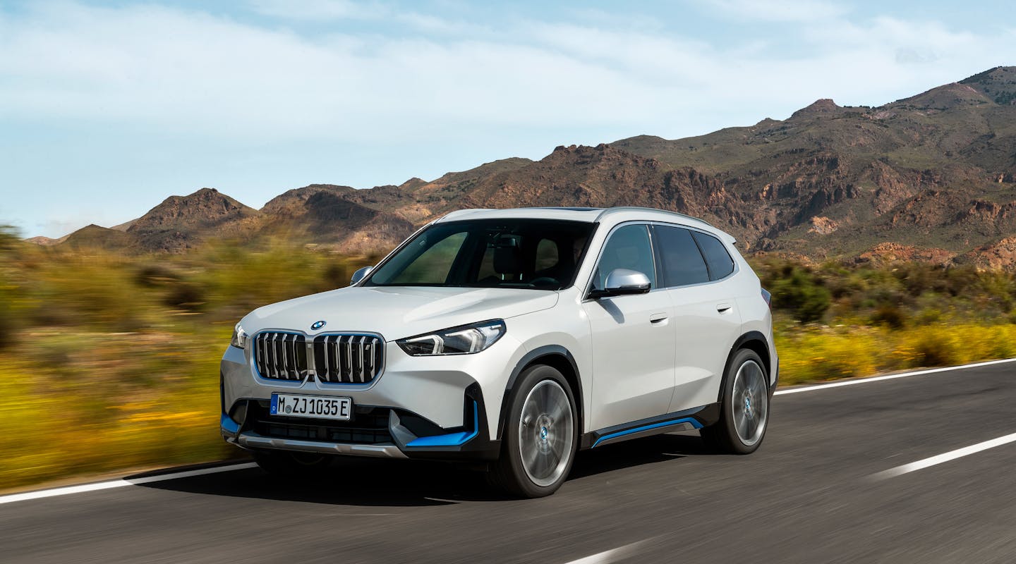 Weißer BMW-SUV fährt auf einer Landstraße durch eine trockene, hügelige Landschaft unter blauem Himmel.