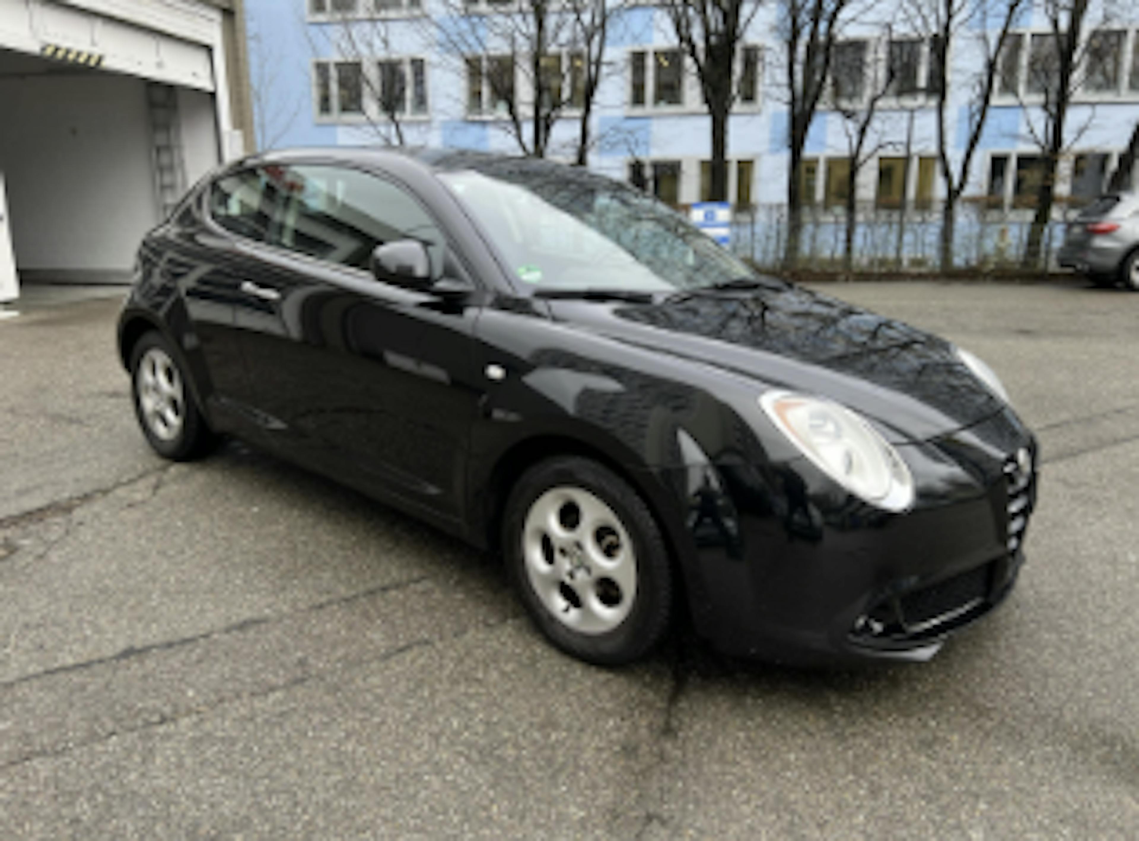 Ein gebrauchter Alfa Romeo MiTo in Schwarz steht auf einem gepflasterten Parkplatz vor einem Gebäude.