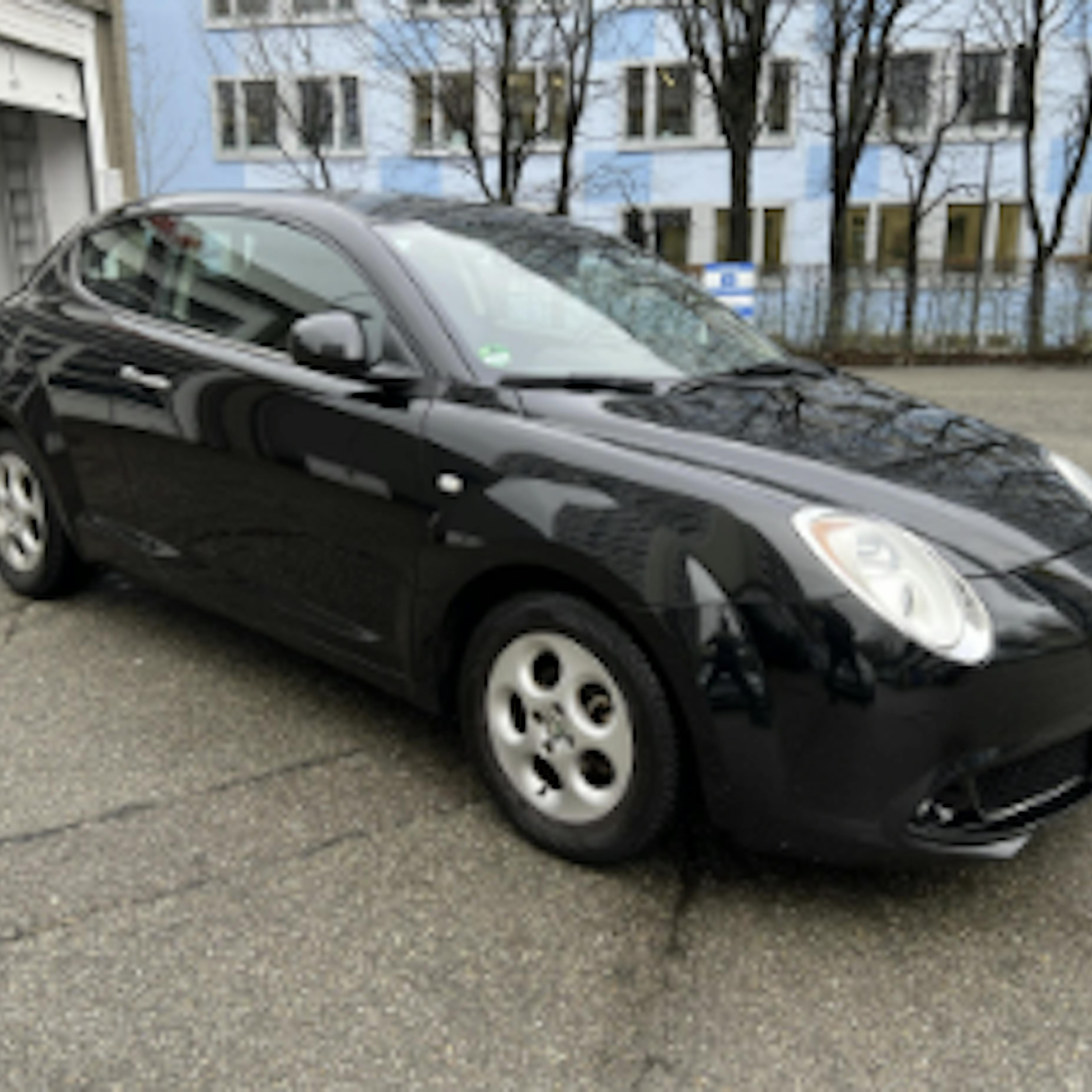 A used Alfa Romeo MiTo in black is parked in a paved parking lot in front of a building.