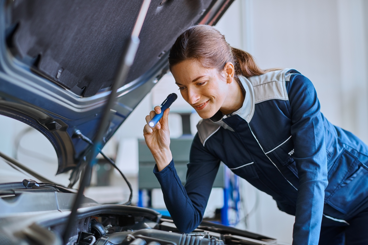 Eine Prüferin des TÜV schaut mit einer Taschenlampe in den Motorraum eines Autos.