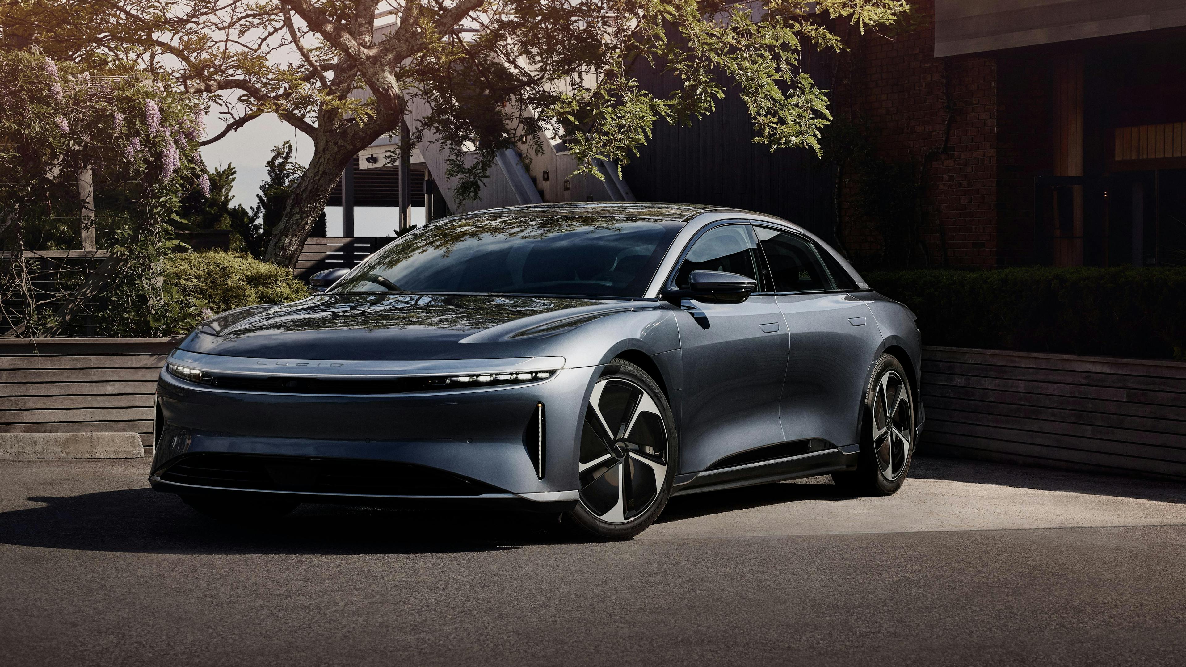 A gray Lucid Air Pure stands in front of a tree and a building.