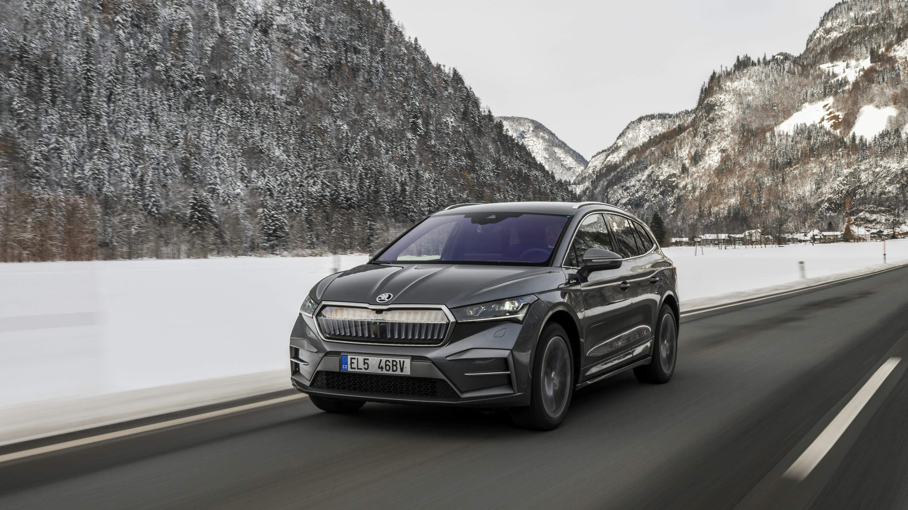 A gray Å koda Enyaq is driving on a country road, snow and mountains in the background.
