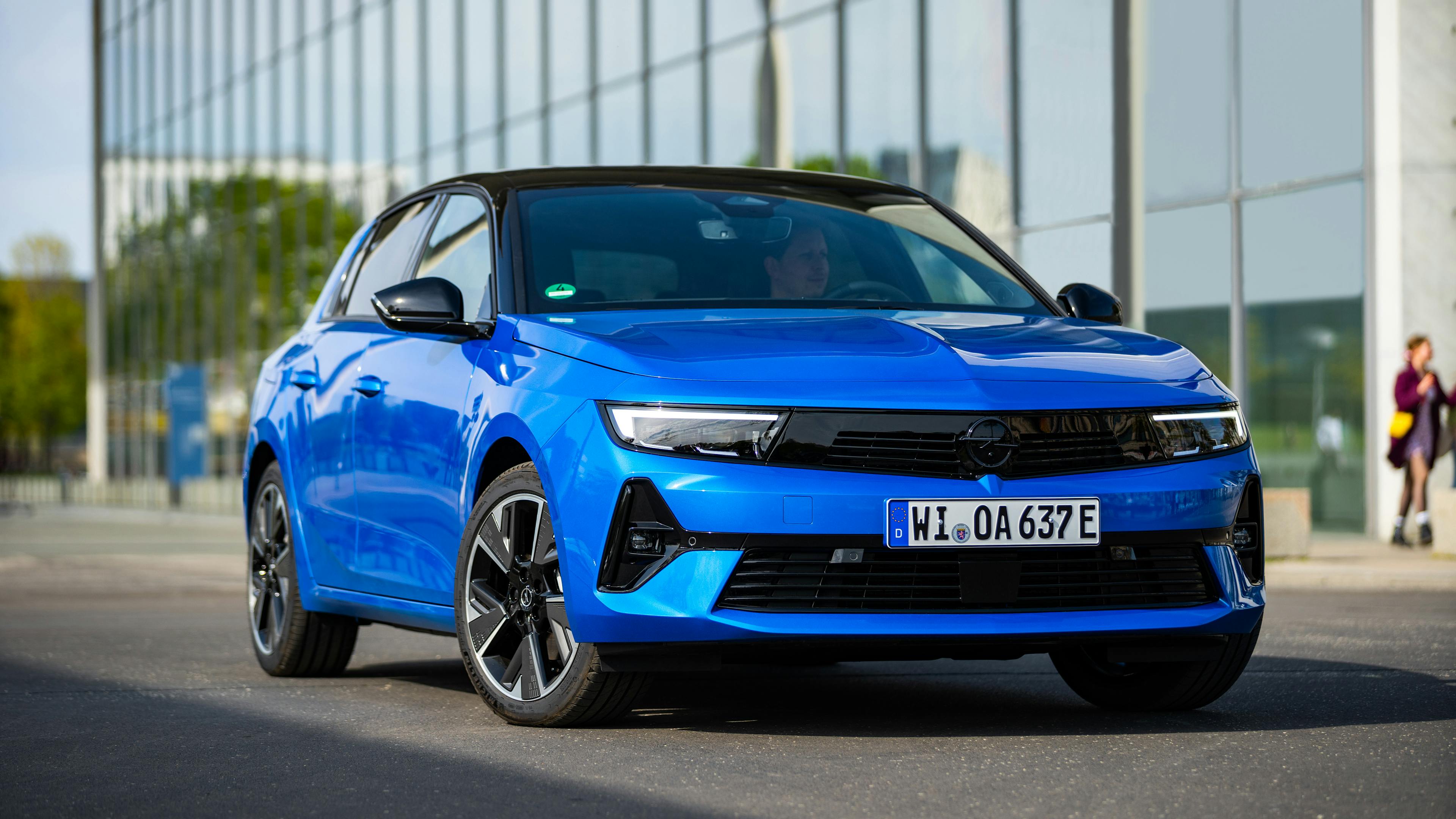 Blue Opel Astra Electric in front of a glazed building wall.
