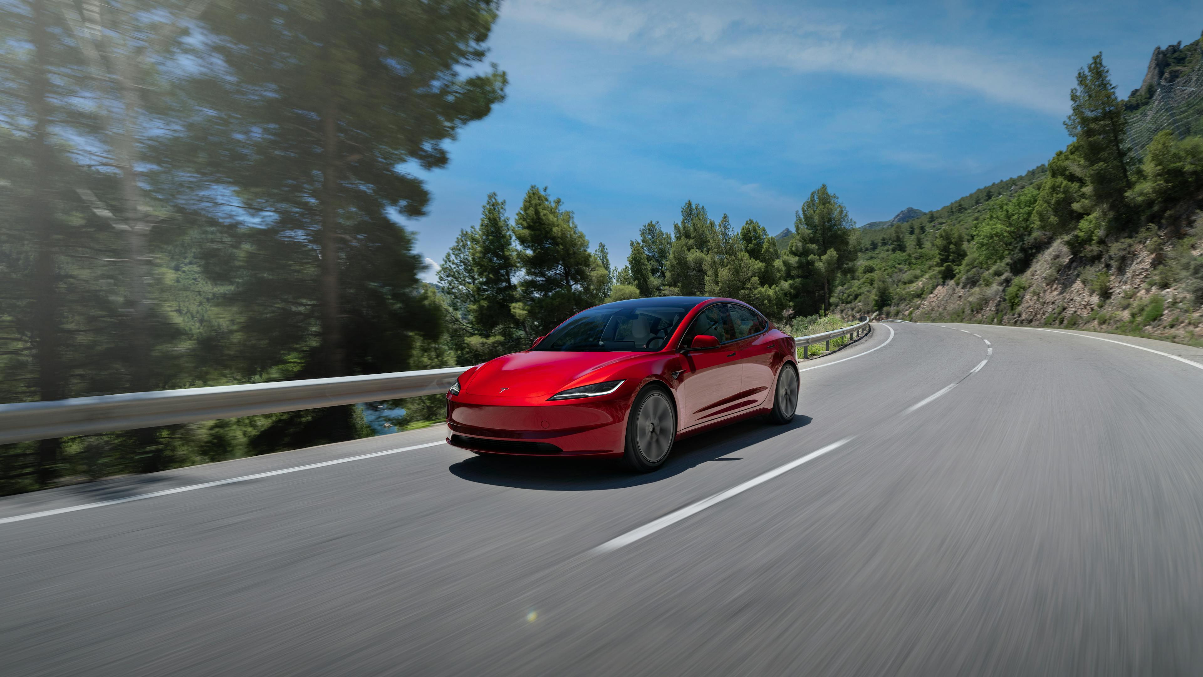 Ein rotes Tesla Model 3 fährt auf einer Landstraße in den Bergen.