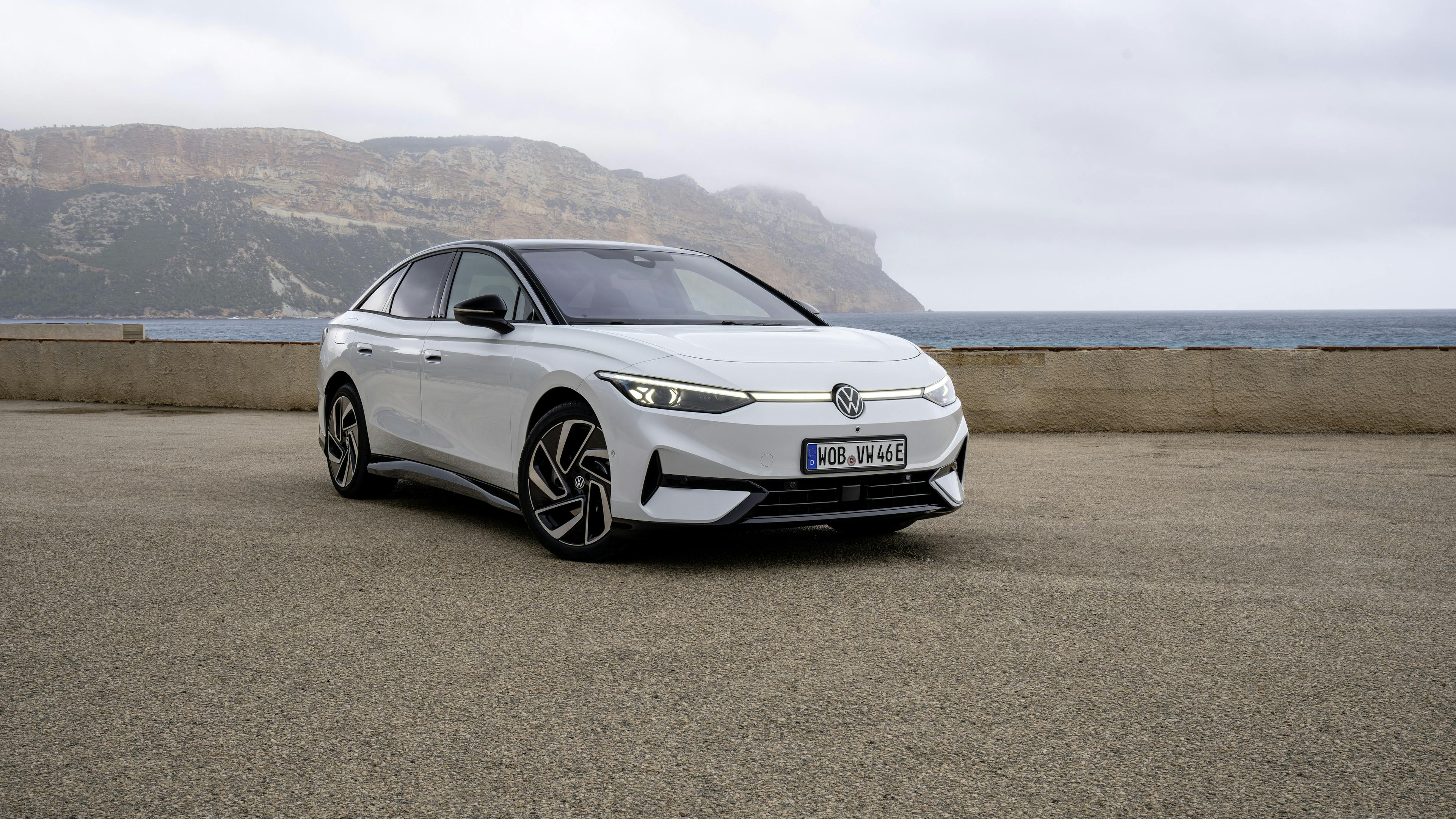 A white VW ID.7 Pro stands in front of a body of water and a mountain.