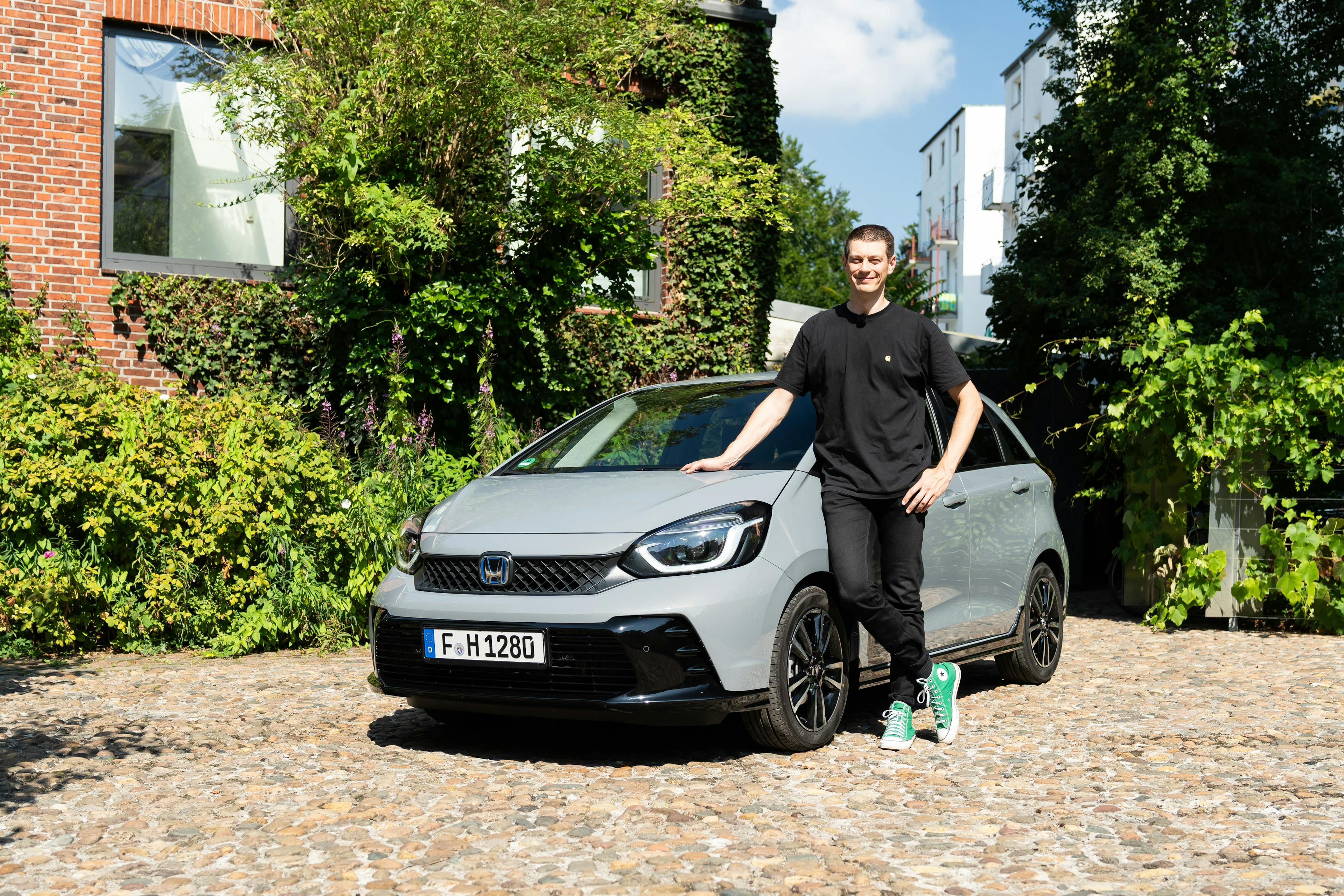 Eine Person lehnt an einem grauen Honda Jazz, der in einer grünen Umgebung mit viel Pflanzenbewuchs im Hintergrund steht.