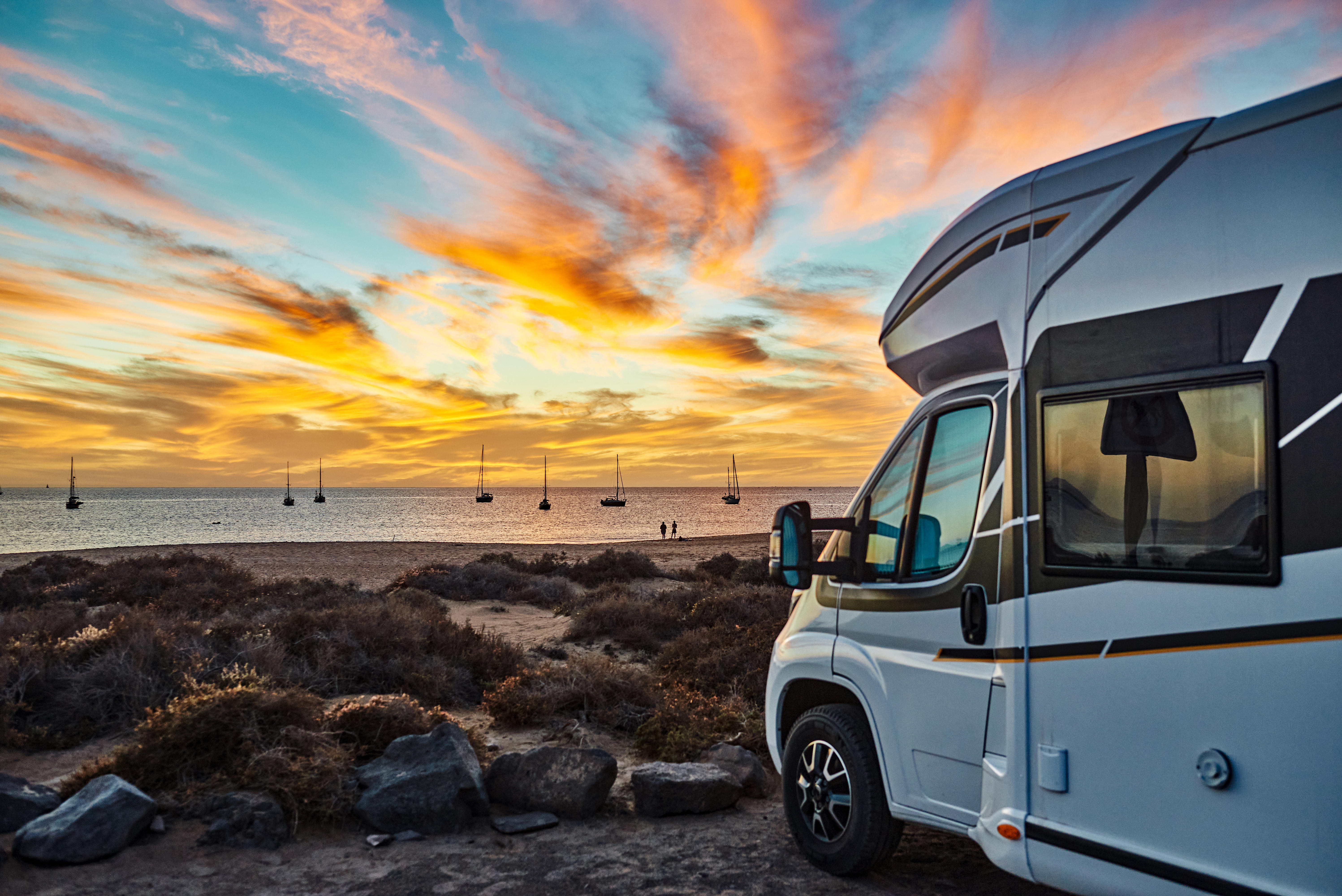 Ein weißes Wohnmobil steht bei Sonnenuntergang am Meer. Auf dem Wasser sind Segelschiffe.