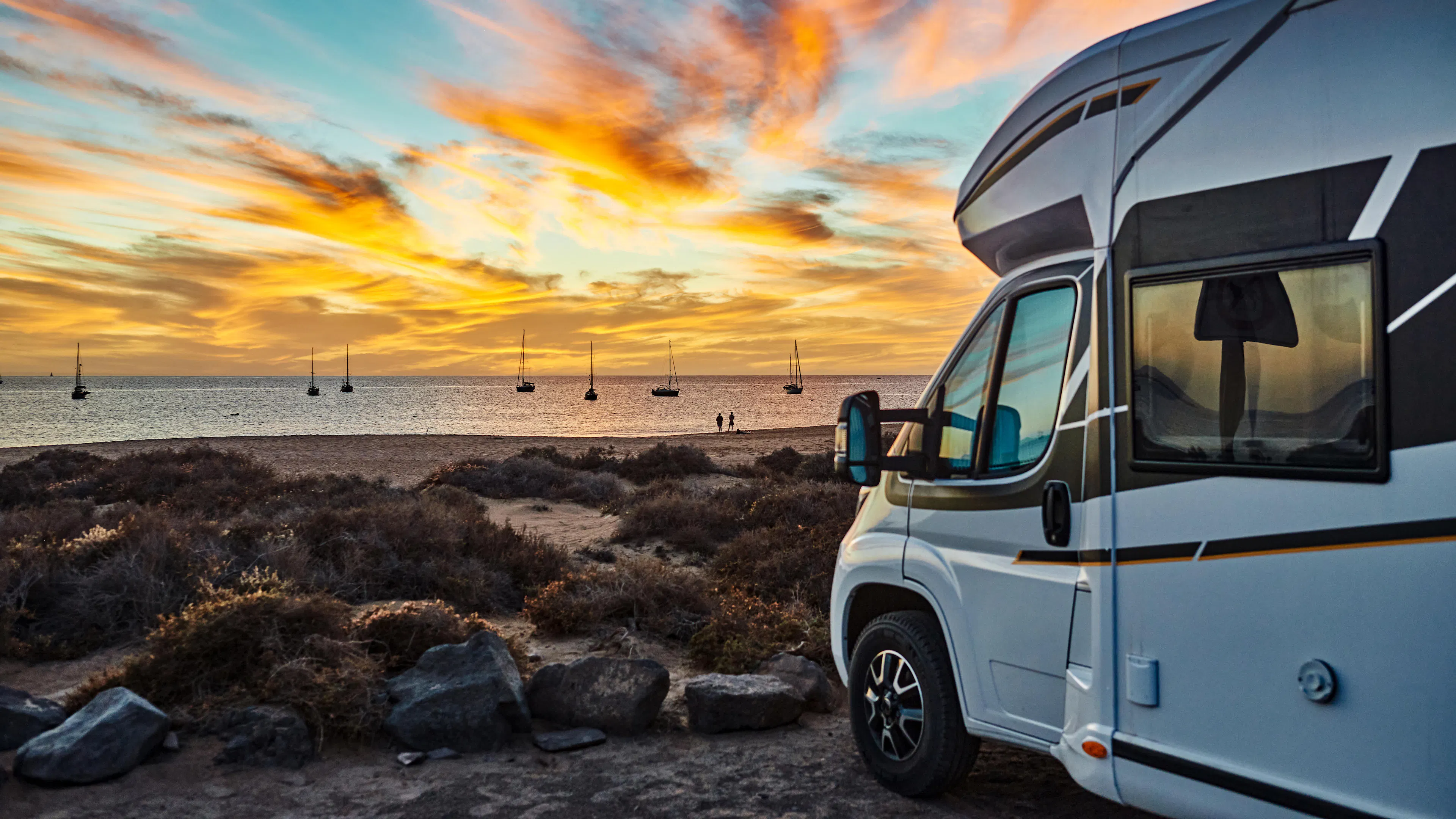 Ein weißes Wohnmobil steht bei Sonnenuntergang am Meer. Auf dem Wasser sind Segelschiffe.