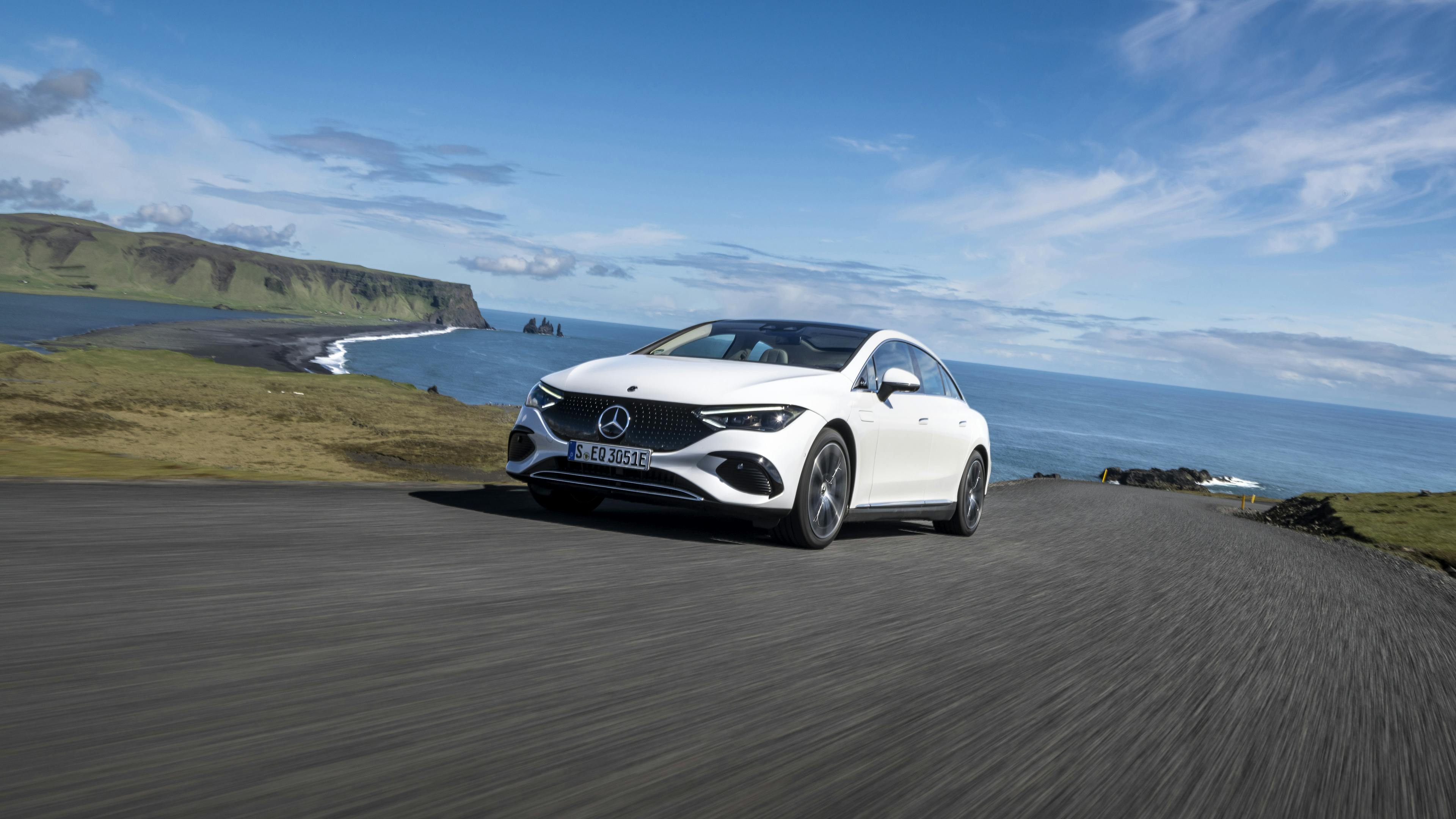 A white Mercedes EQE is driving on a coastal road with a cliff in the background.