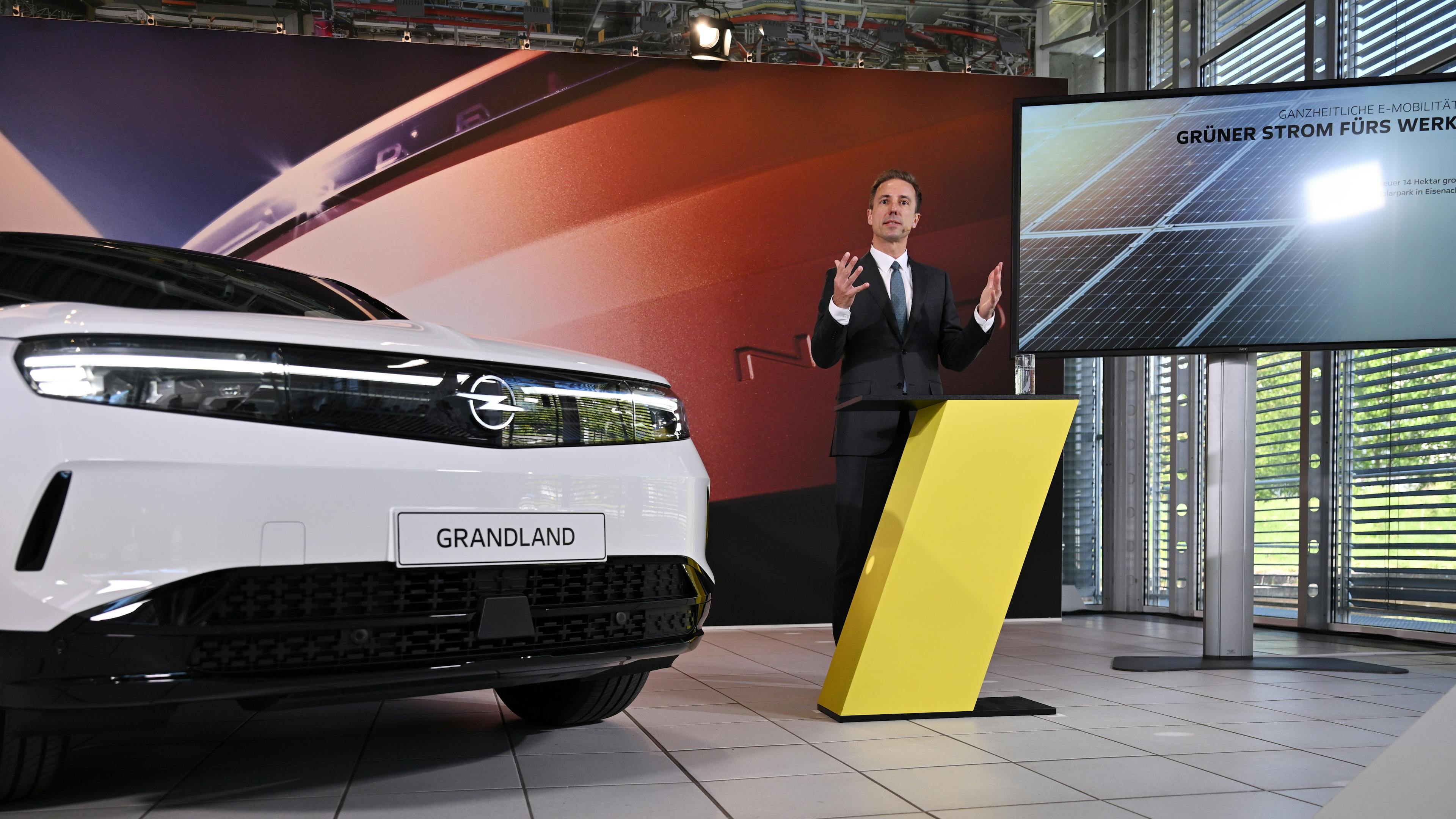 Opel CEO Florian Huettl stands gesticulating next to a white Opel Grandland during a presentation.
