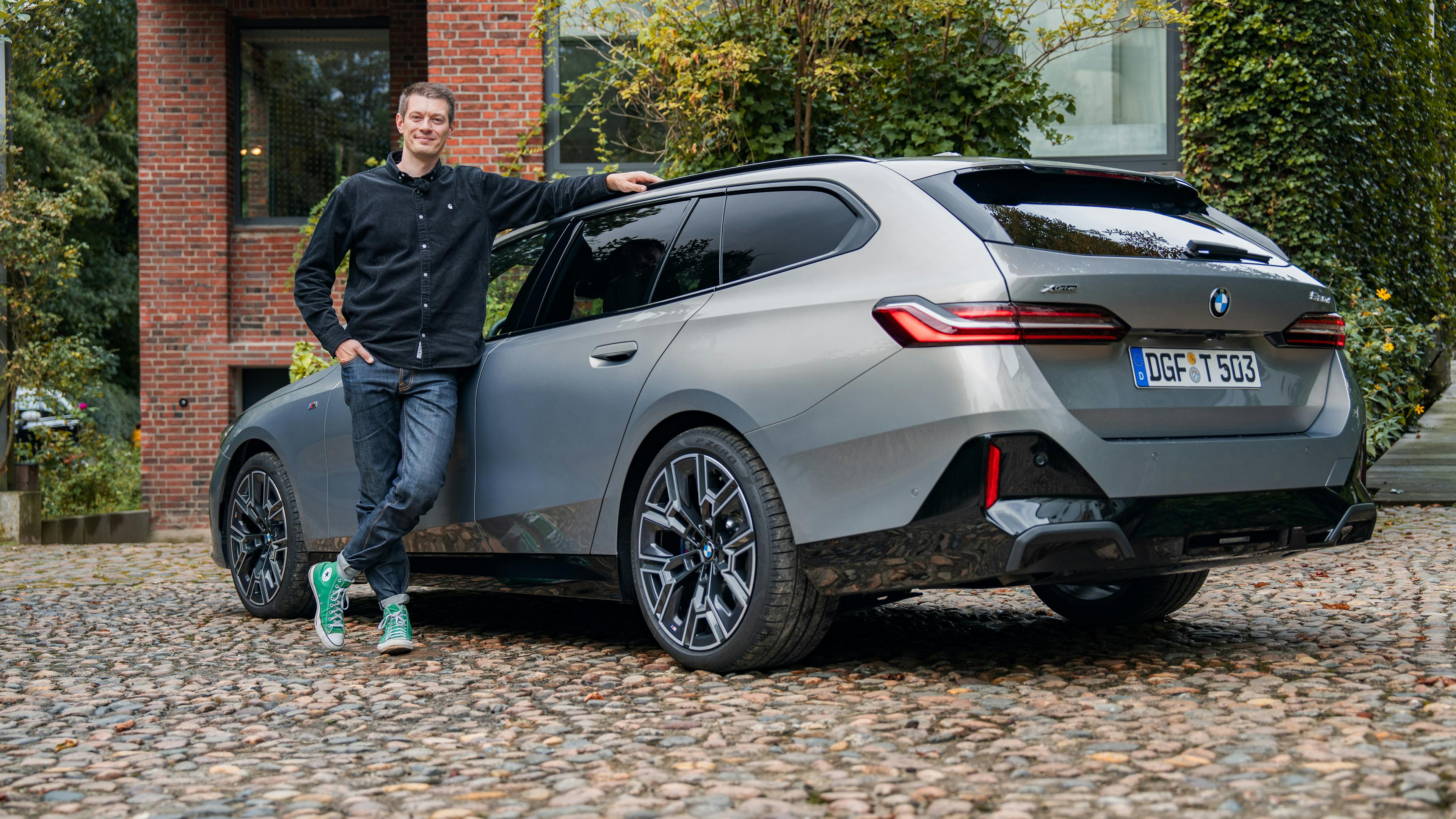  Silberner BMW 520d xDrive Touring mit M-Sportpaket auf Kopfsteinpflasterplatz mit Klinkergebäude im Hintergrund und dem Redakteur am Auto lehnend.