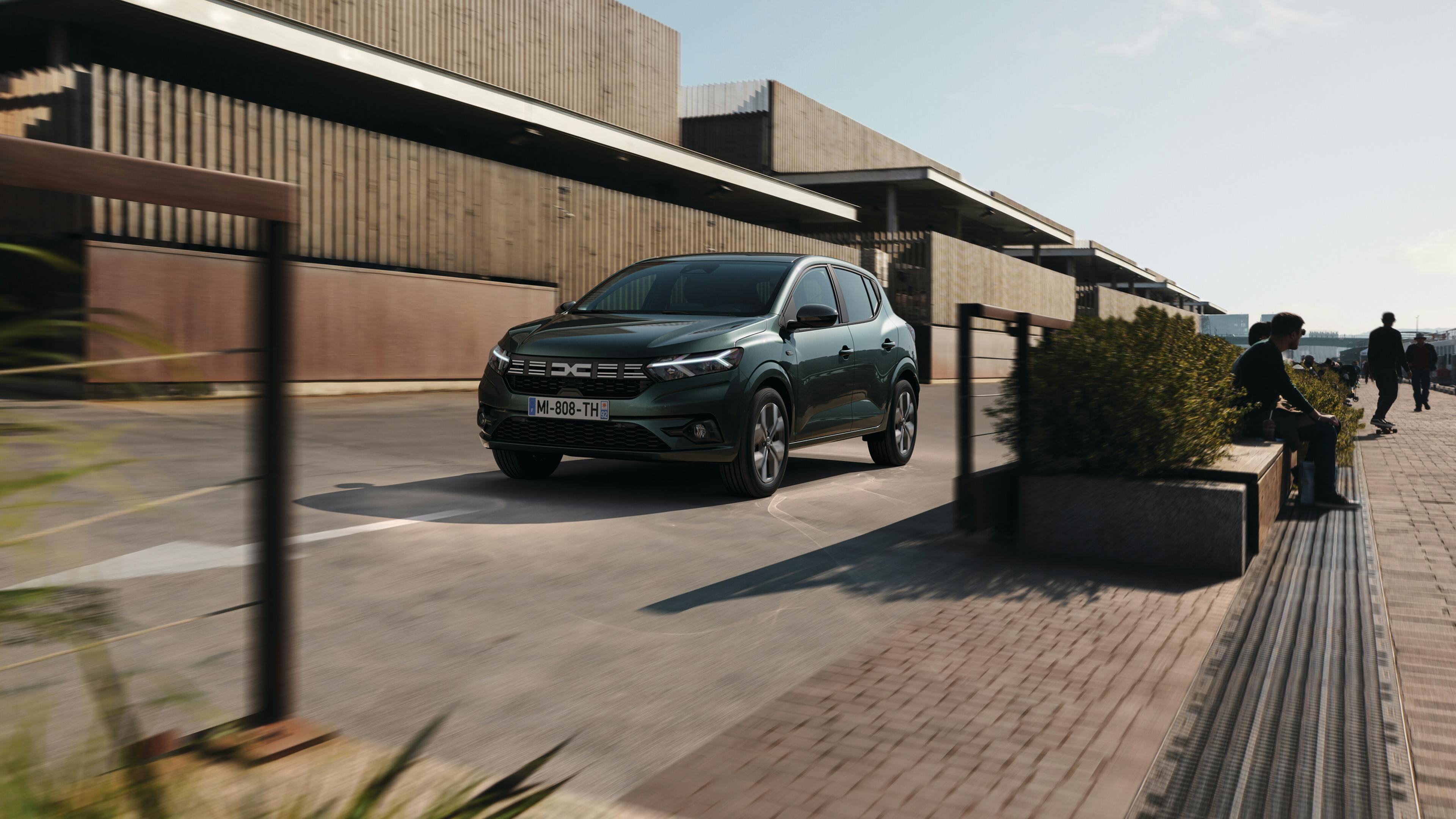 Ein dunkelgrüner Dacia Sandero fährt eine Straße neben modernen, minimalistischen Gebäuden entlang.