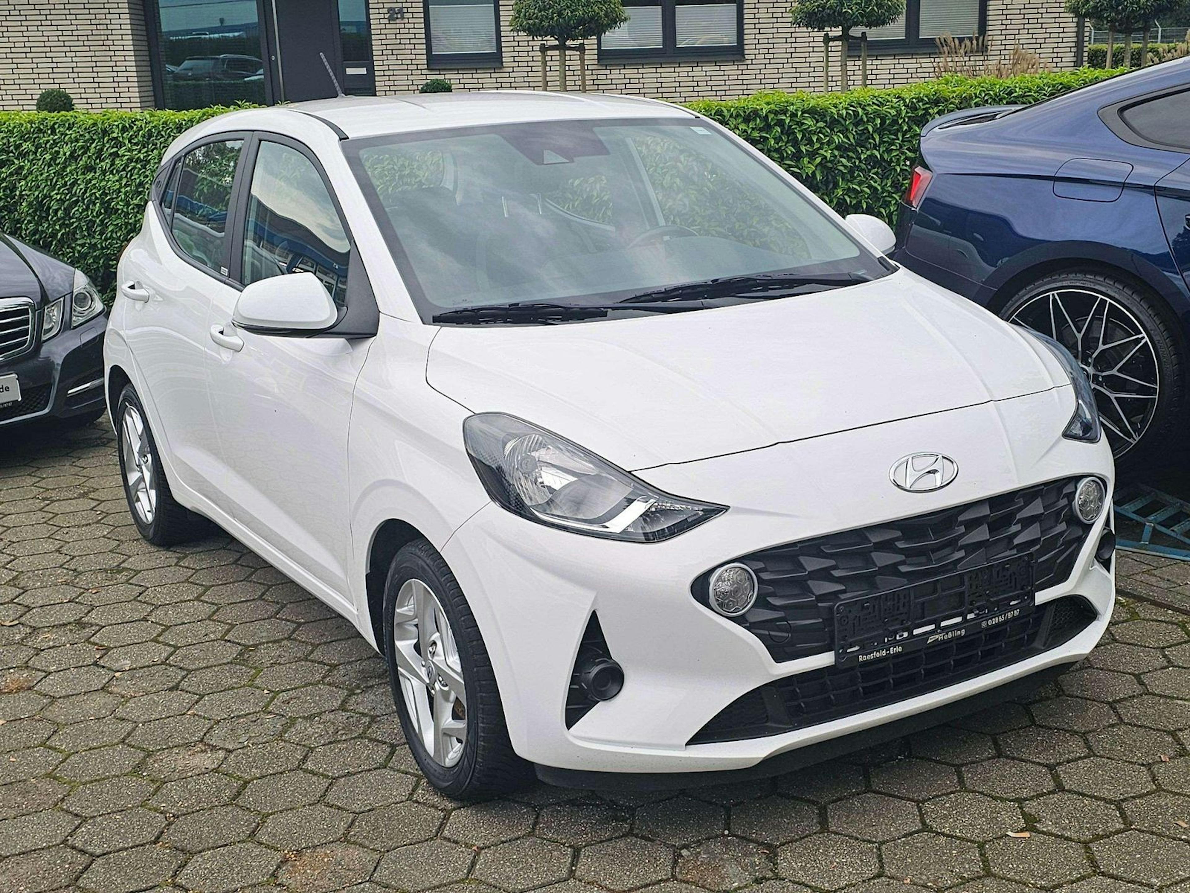 Ein weißer Hyundai i10 steht auf einem gepflasterten Parkplatz neben anderen Autos.