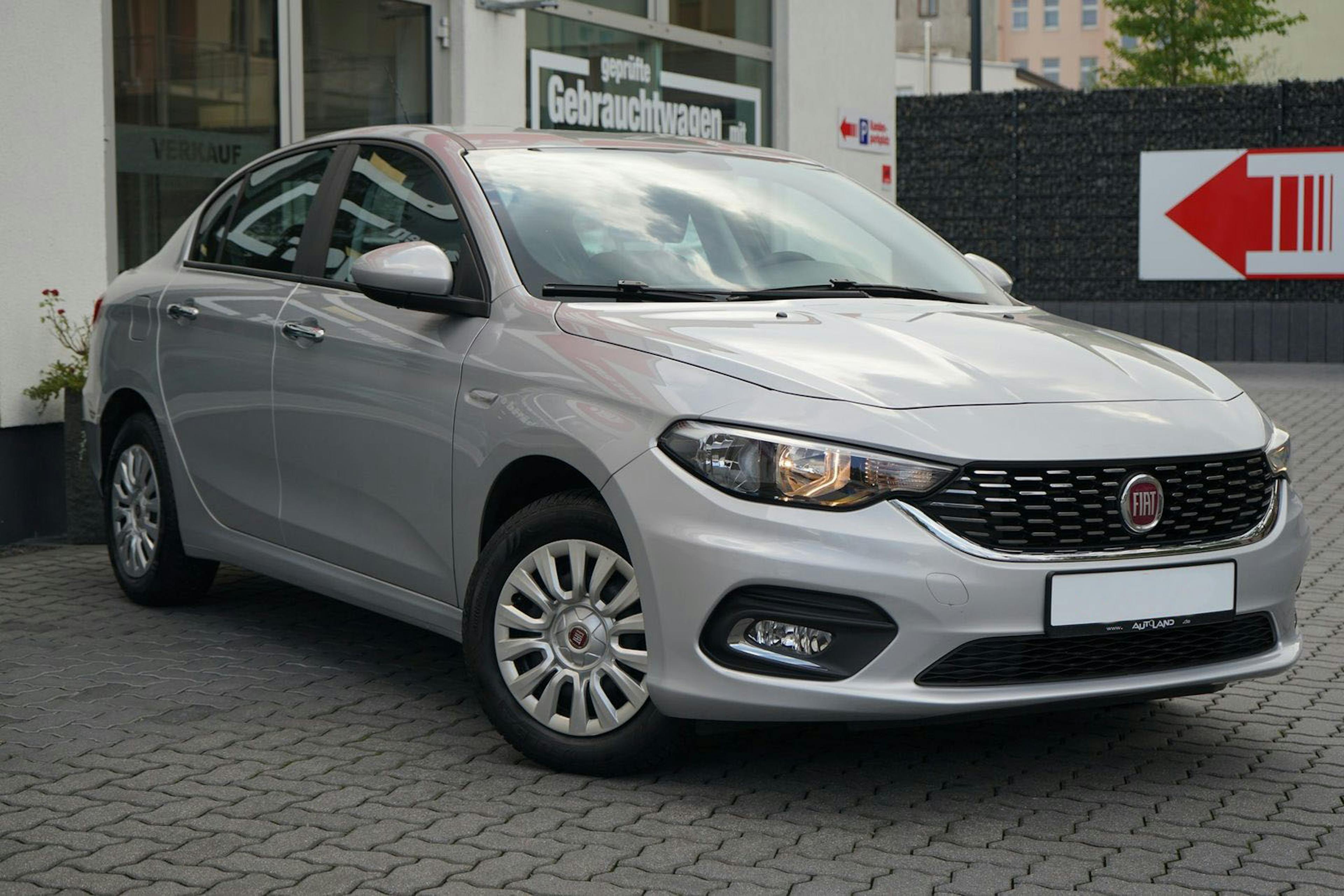 Ein silberfarbener Fiat Tipo parkt vor einem Autohaus auf einem gepflasterten Boden.