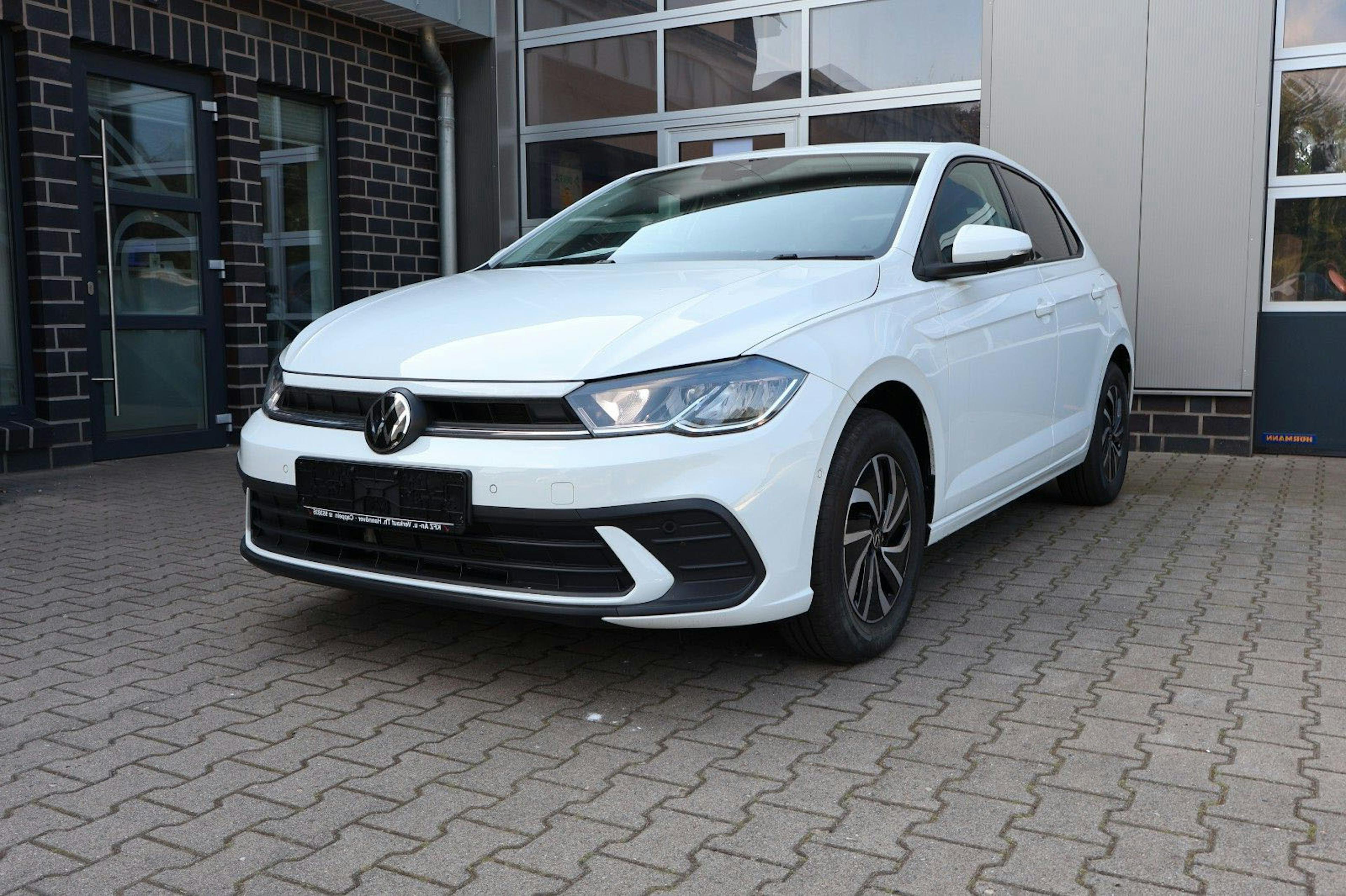 Ein weißer VW Polo parkt vor einem Autohaus auf einem gepflasterten Parkplatz.