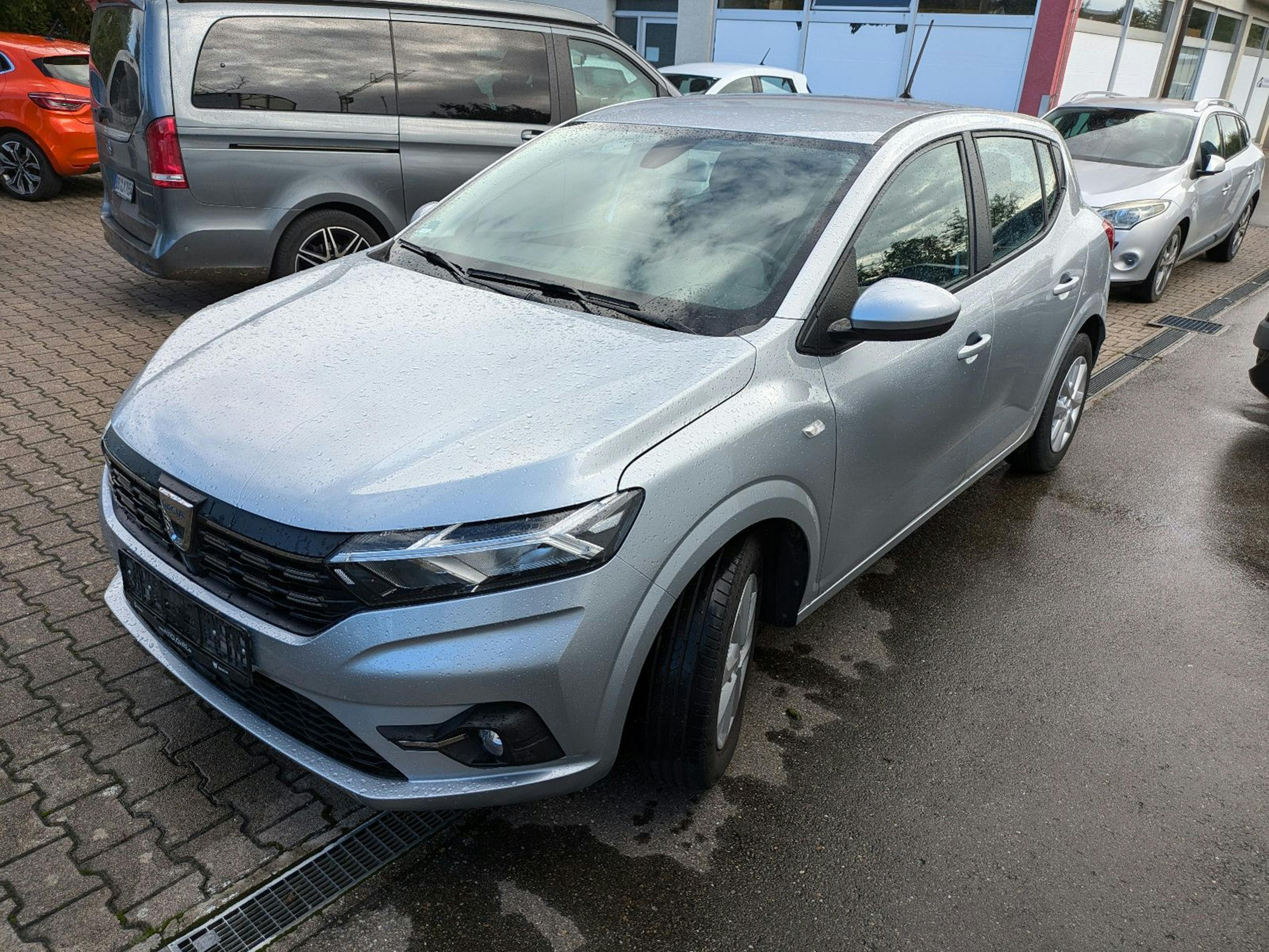 Ein silberfarbener Dacia Sandero steht auf einem Parkplatz neben anderen Fahrzeugen.