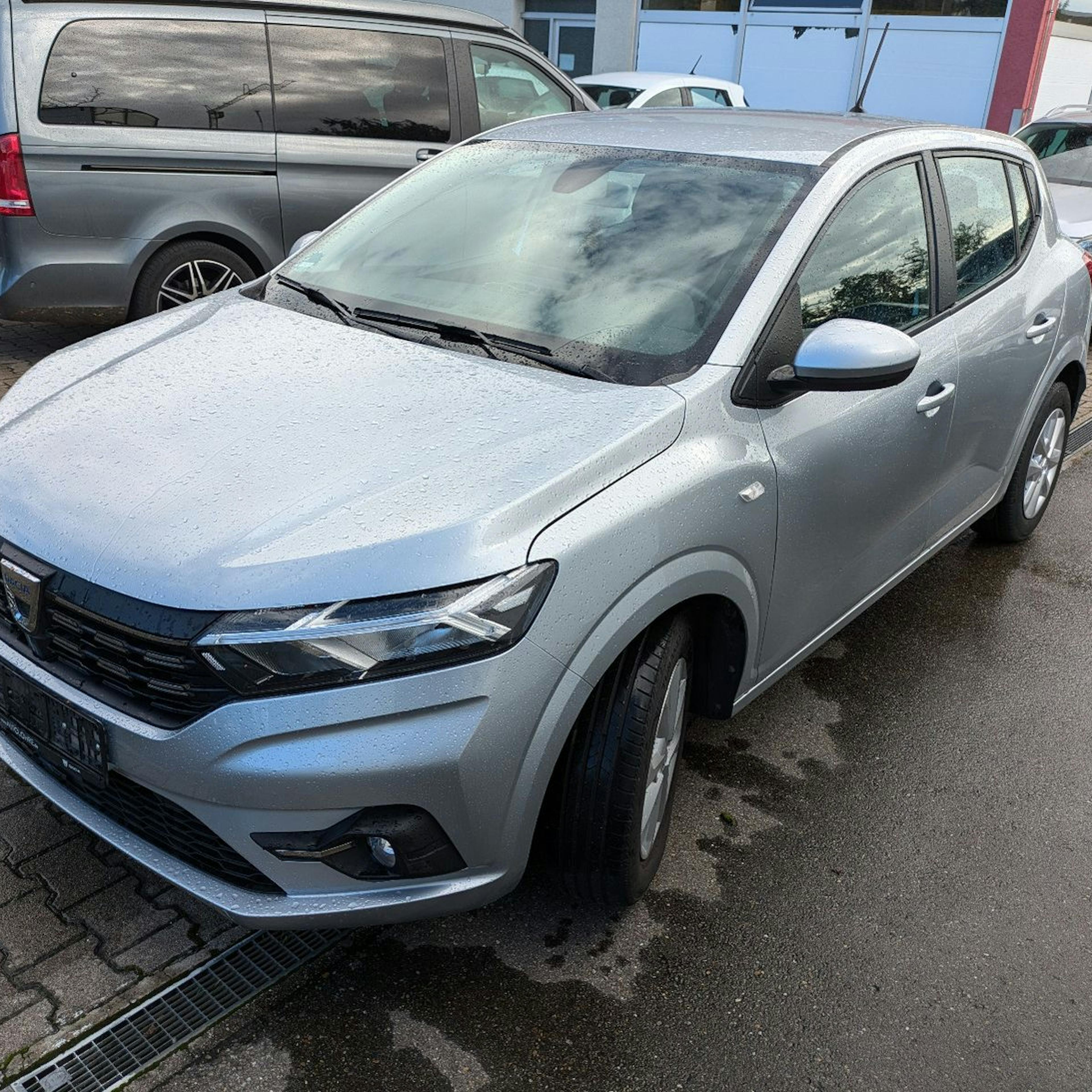 A silver-colored Dacia Sandero is parked in a parking lot next to other vehicles.