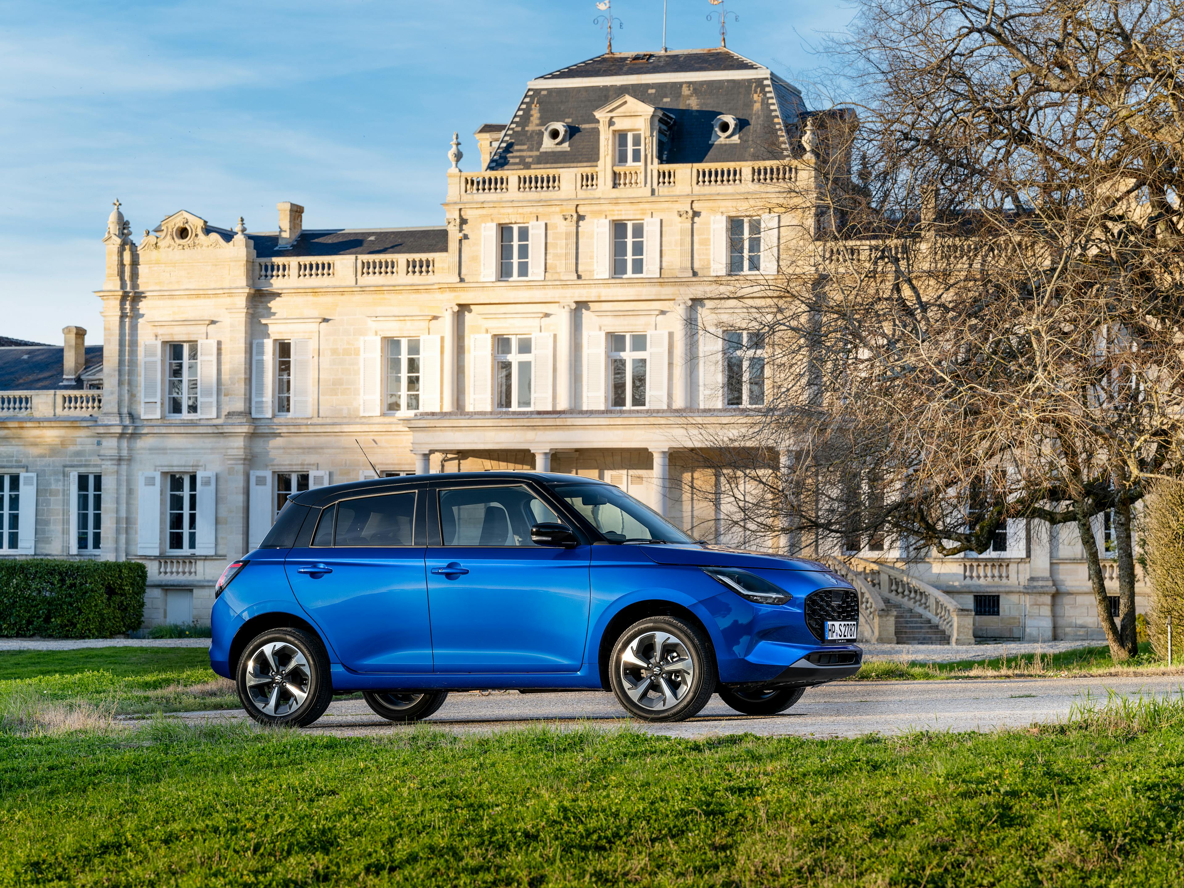 Ein blauer Suzuki Swift steht vor einem klassischen Herrenhaus in einer ländlichen Umgebung.