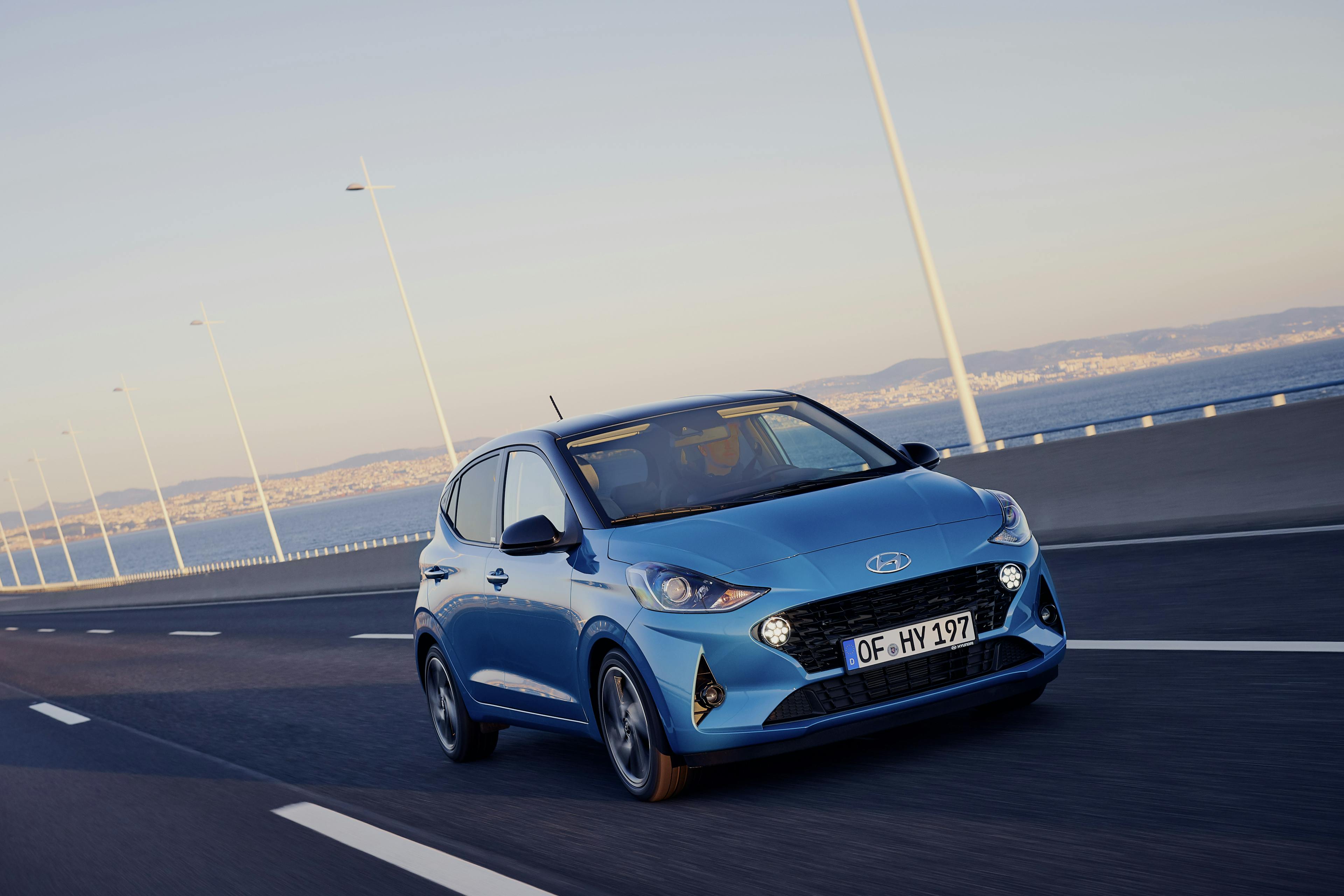 Ein blauer Hyundai i10 fährt auf einer Straße mit einem weiten Blick auf eine Küste im Hintergrund.